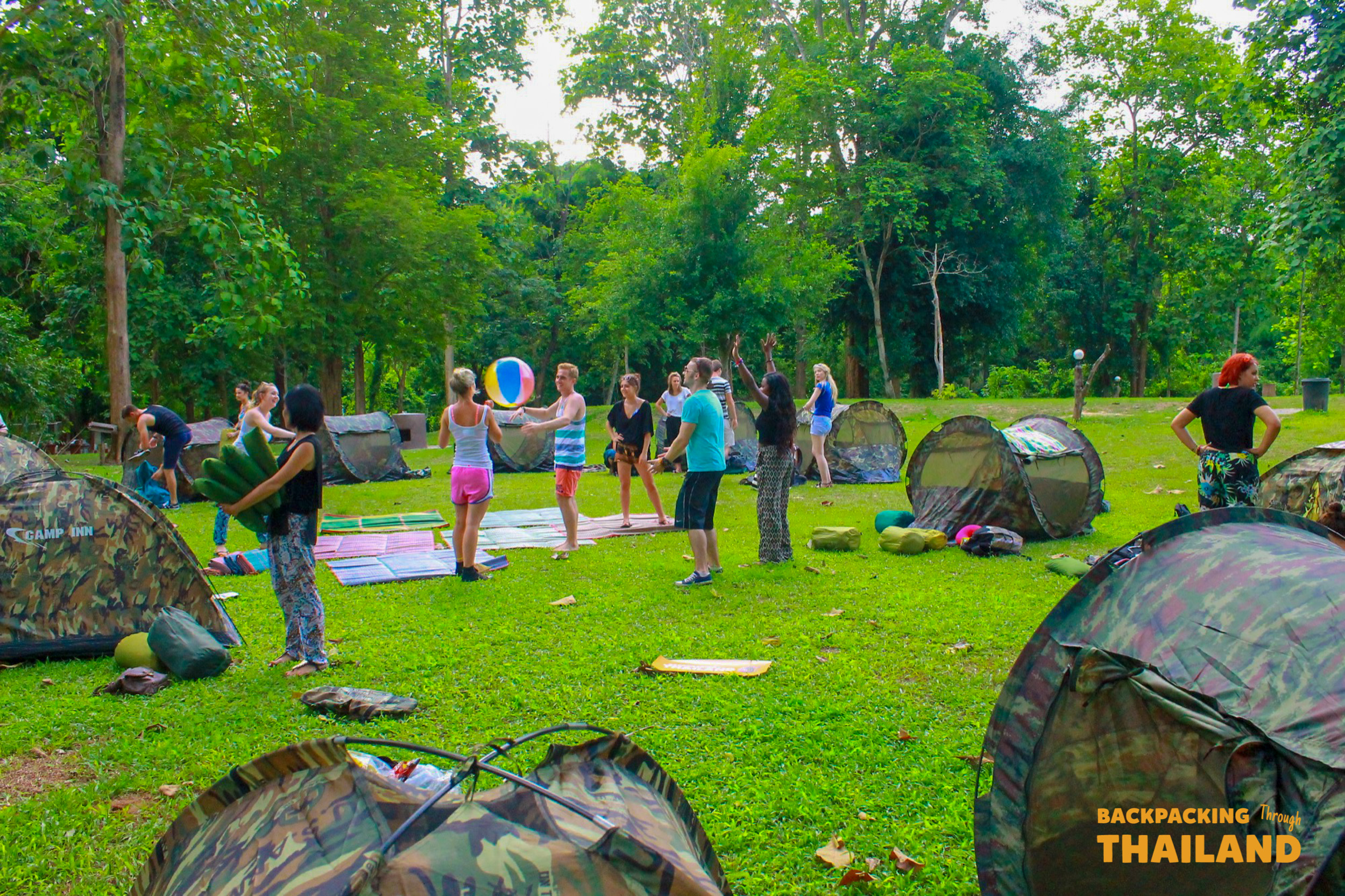 Backpackers playing volleyball and camping in a lush green clearing at Erawan National Park, Thailand.