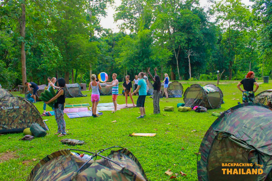 Backpackers playing volleyball and camping in a lush green clearing at Erawan National Park, Thailand.