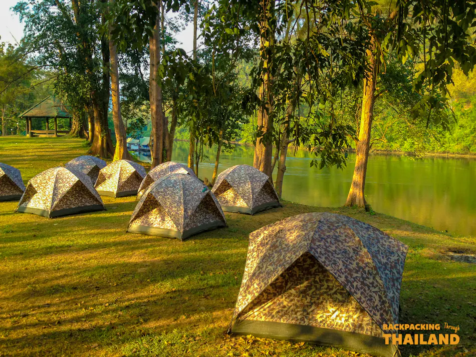 Camouflage tents pitched on green grass beside a calm lake in Erawan National Park, Thailand.