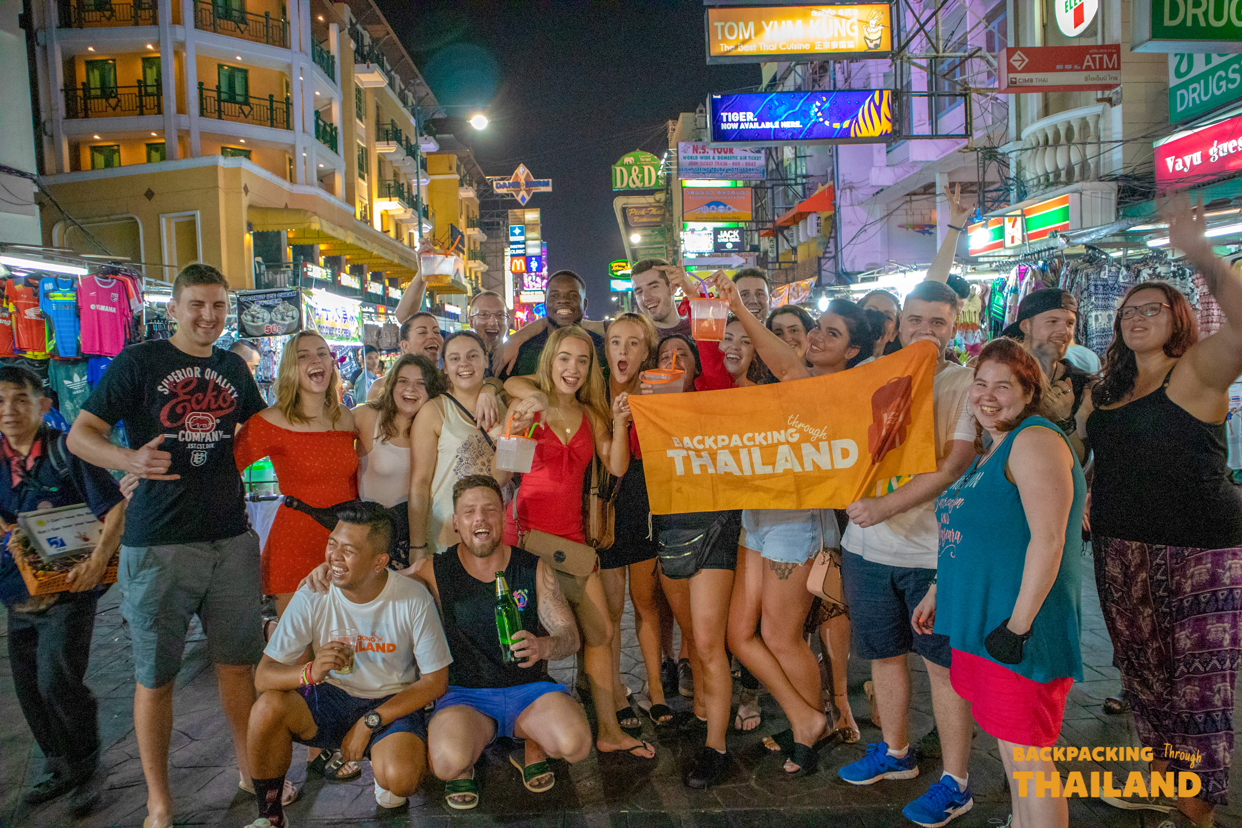 Excited backpackers pose with a 'Backpackers Thailand' flag on a lively Khao San Road at night.