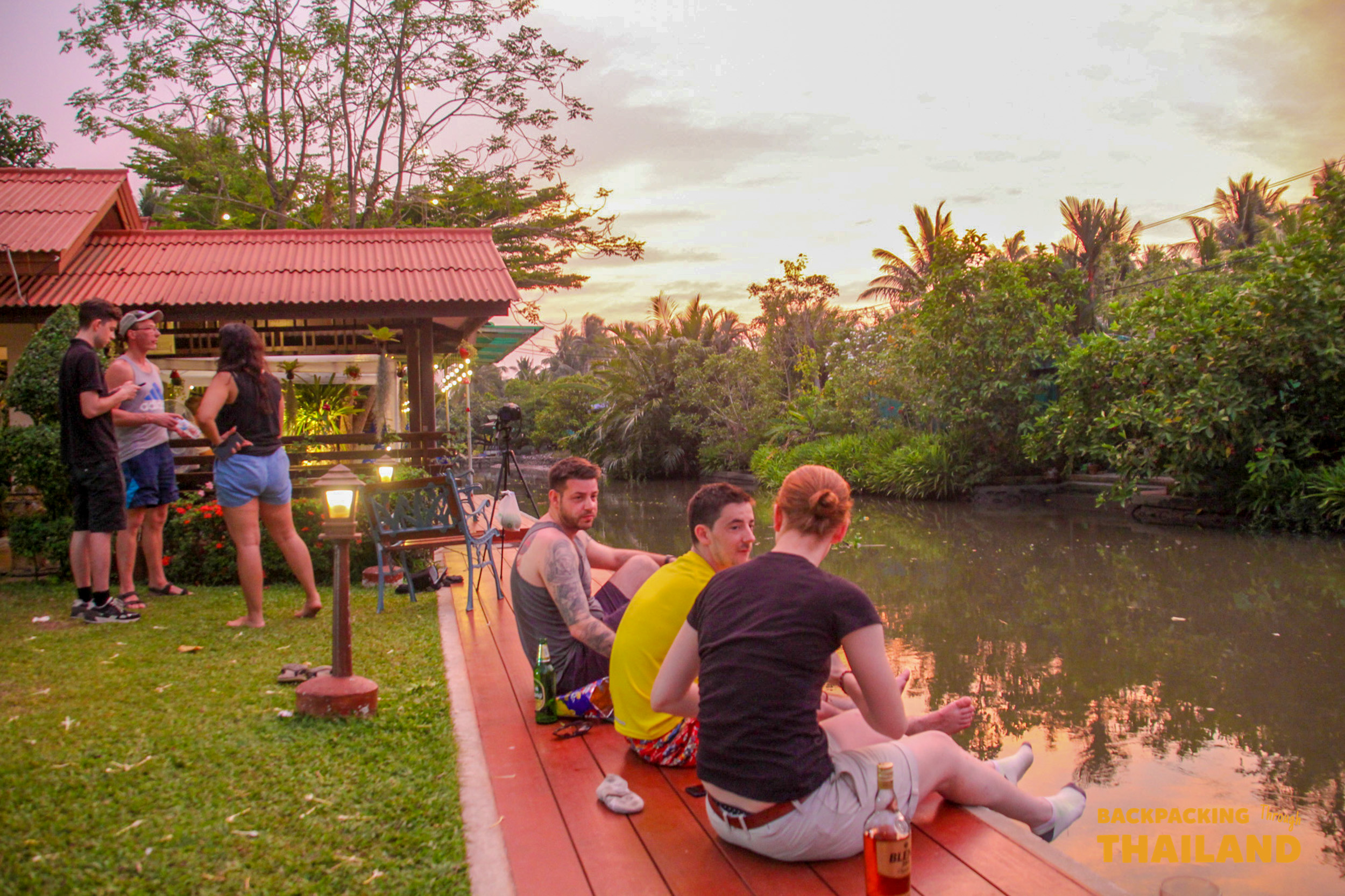 Travelers relax by a canal at a homestay in Amphawa, Thailand, enjoying the evening sunset.