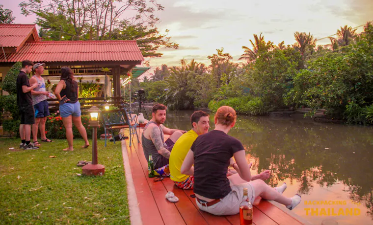 Travelers relax by a canal at a homestay in Amphawa, Thailand, enjoying the evening sunset.