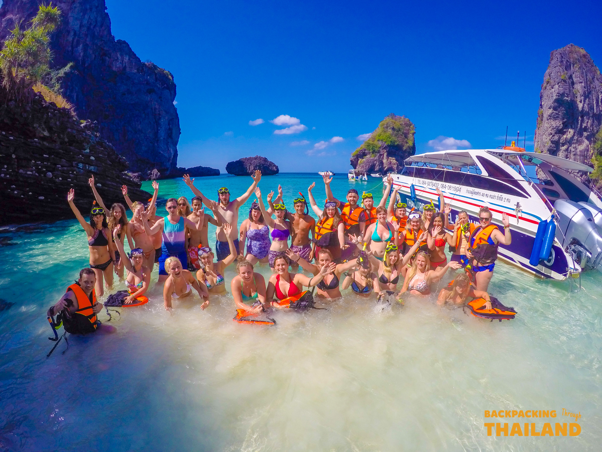 A large group of snorkellers in life jackets stand waist-deep in turquoise water beside their speedboat, arms raised, below the limestone cliffs of Koh Phi Phi.