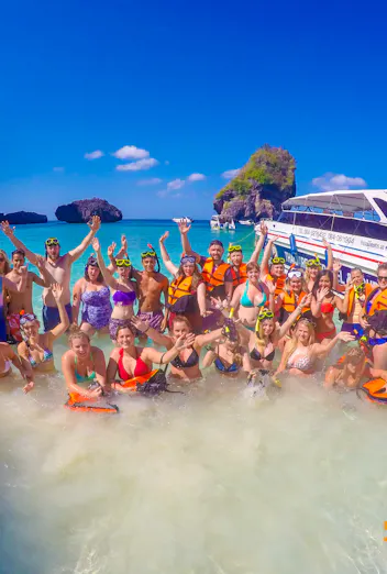 A large group of snorkellers in life jackets stand waist-deep in turquoise water beside their speedboat, arms raised, below the limestone cliffs of Koh Phi Phi.