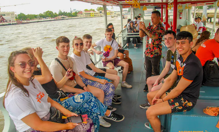 Smiling backpackers on a river boat tour in Bangkok, Thailand, listening to their guide.