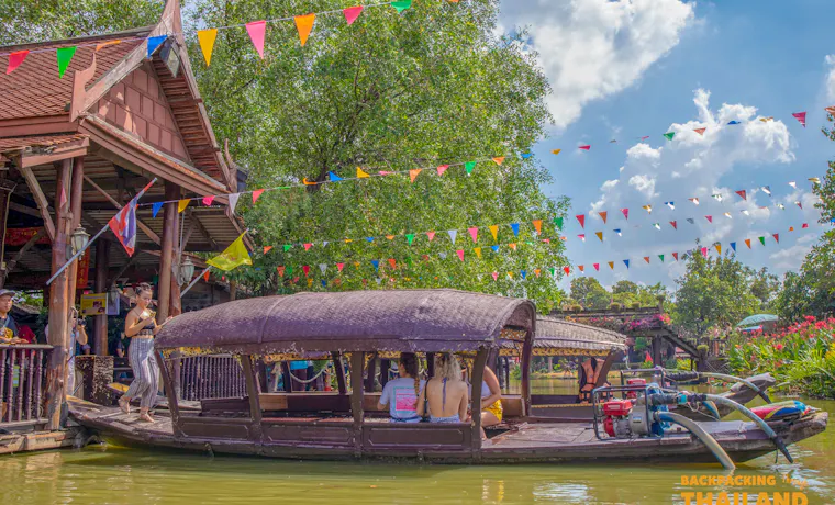 Visitors enjoy a scenic boat ride past traditional Thai buildings and festive flags under a blue sky.
