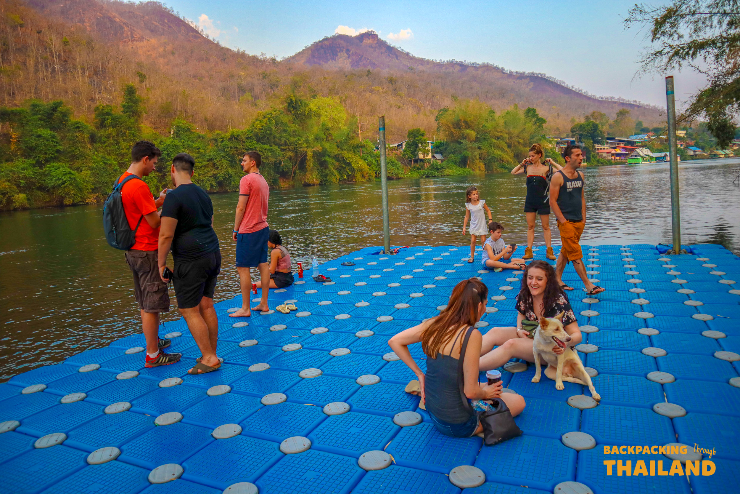 Backpackers and a dog relax on a blue floating platform on the Khwae Yai River.