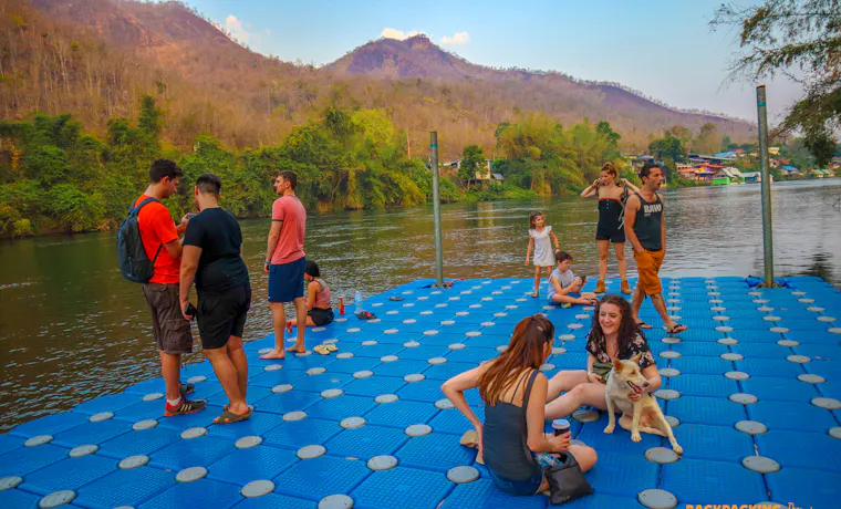 Backpackers and a dog relax on a blue floating platform on the Khwae Yai River.