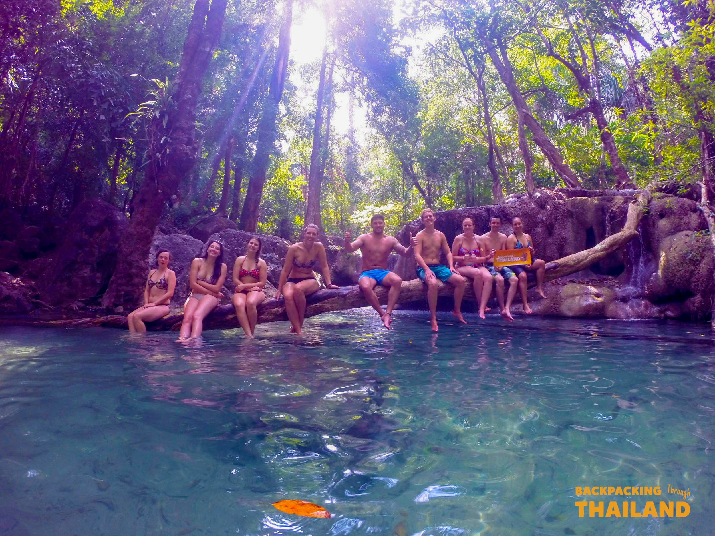 A group of happy backpackers relax on a log over clear emerald water at Erawan Waterfalls, Thailand.