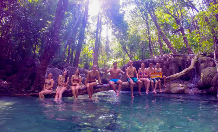 A group of happy backpackers relax on a log over clear emerald water at Erawan Waterfalls, Thailand.