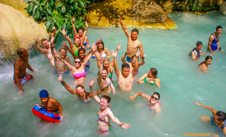 Joyful backpackers raise their arms while swimming in the refreshing emerald waters of Erawan National Park.
