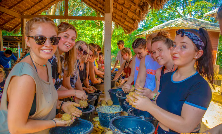 Smiling backpackers prepare food for elephants in buckets at a sunny Thai nature park.