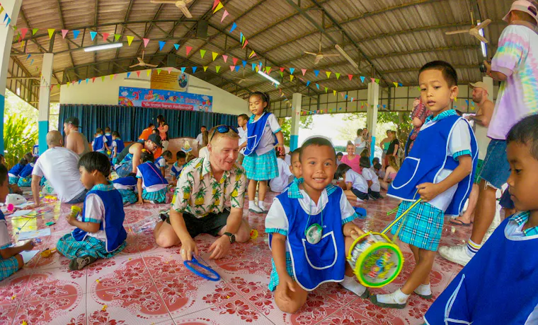 A backpacker plays with smiling Thai school children on a tiled floor during a heartwarming visit in Thailand.