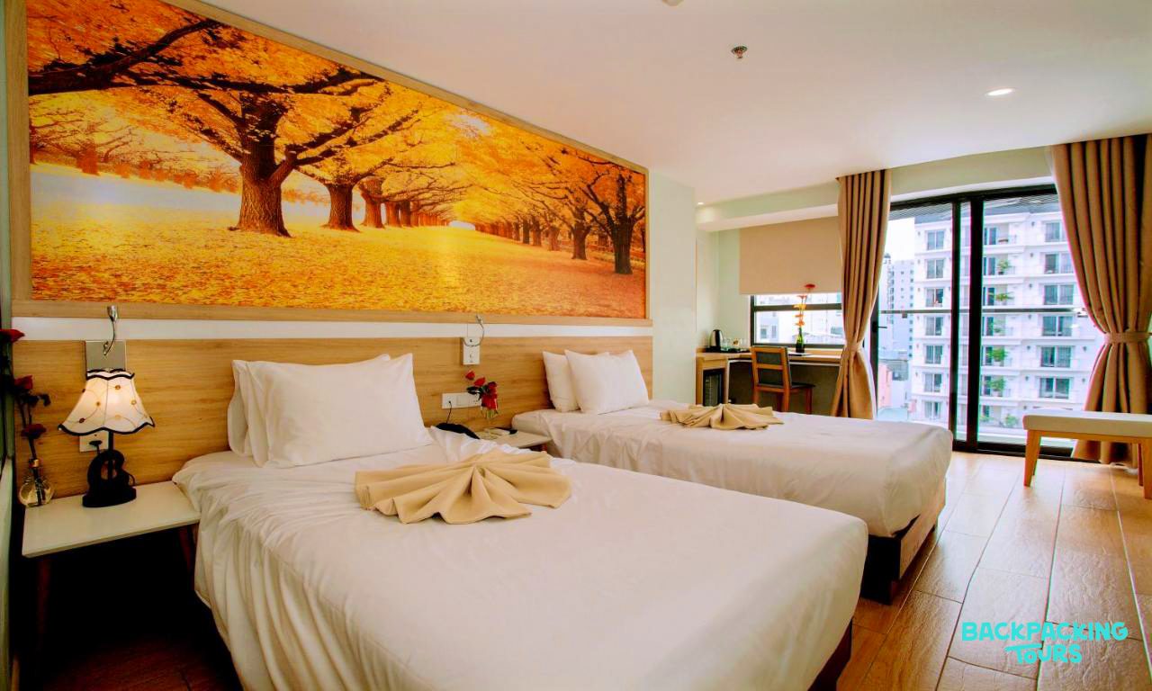 A bright hotel room in Nha Trang featuring two single beds, an autumn mural, and city views.