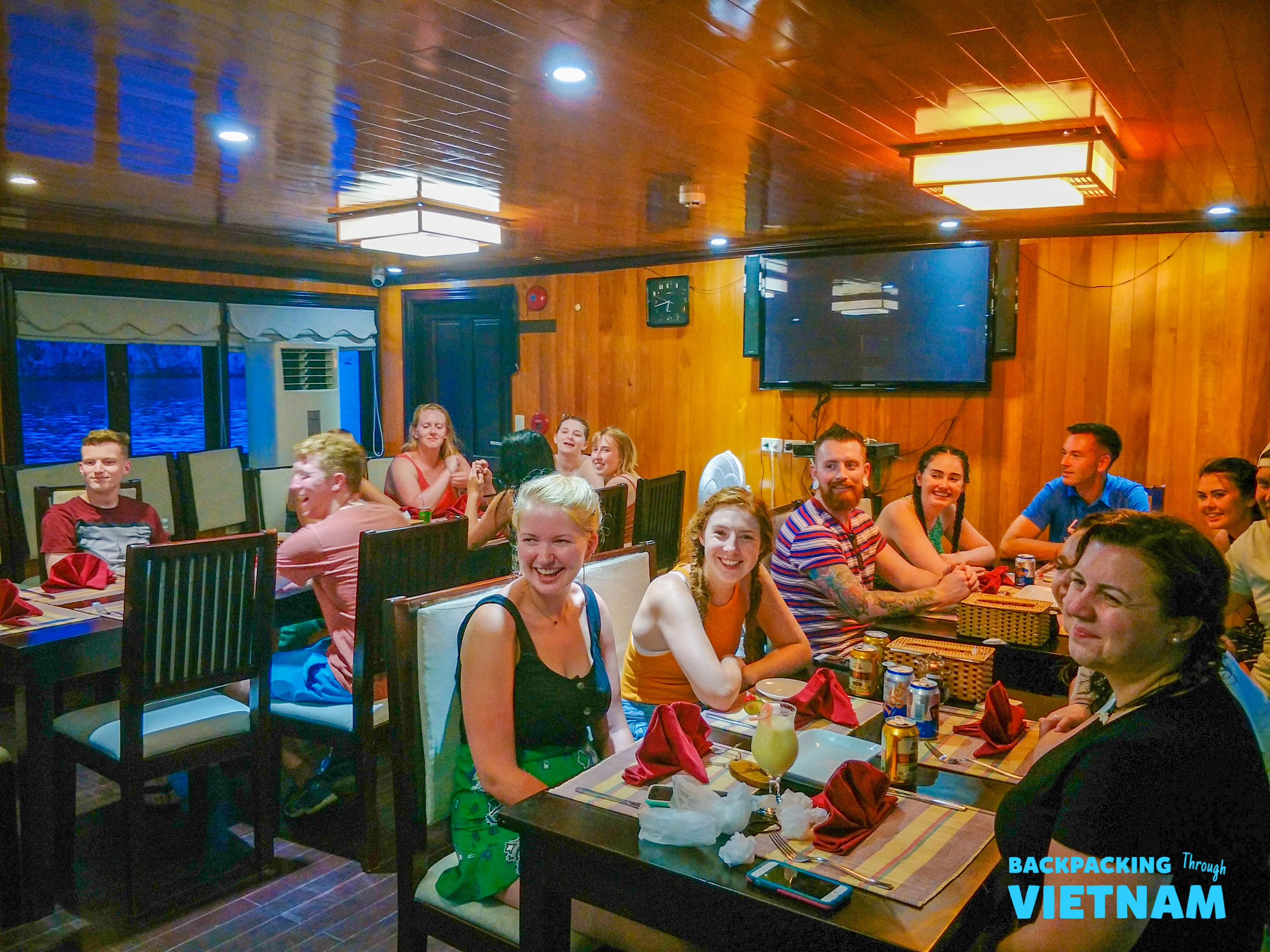 Smiling backpackers enjoy dinner and drinks inside a cozy wooden dining room on their Halong Bay boat.