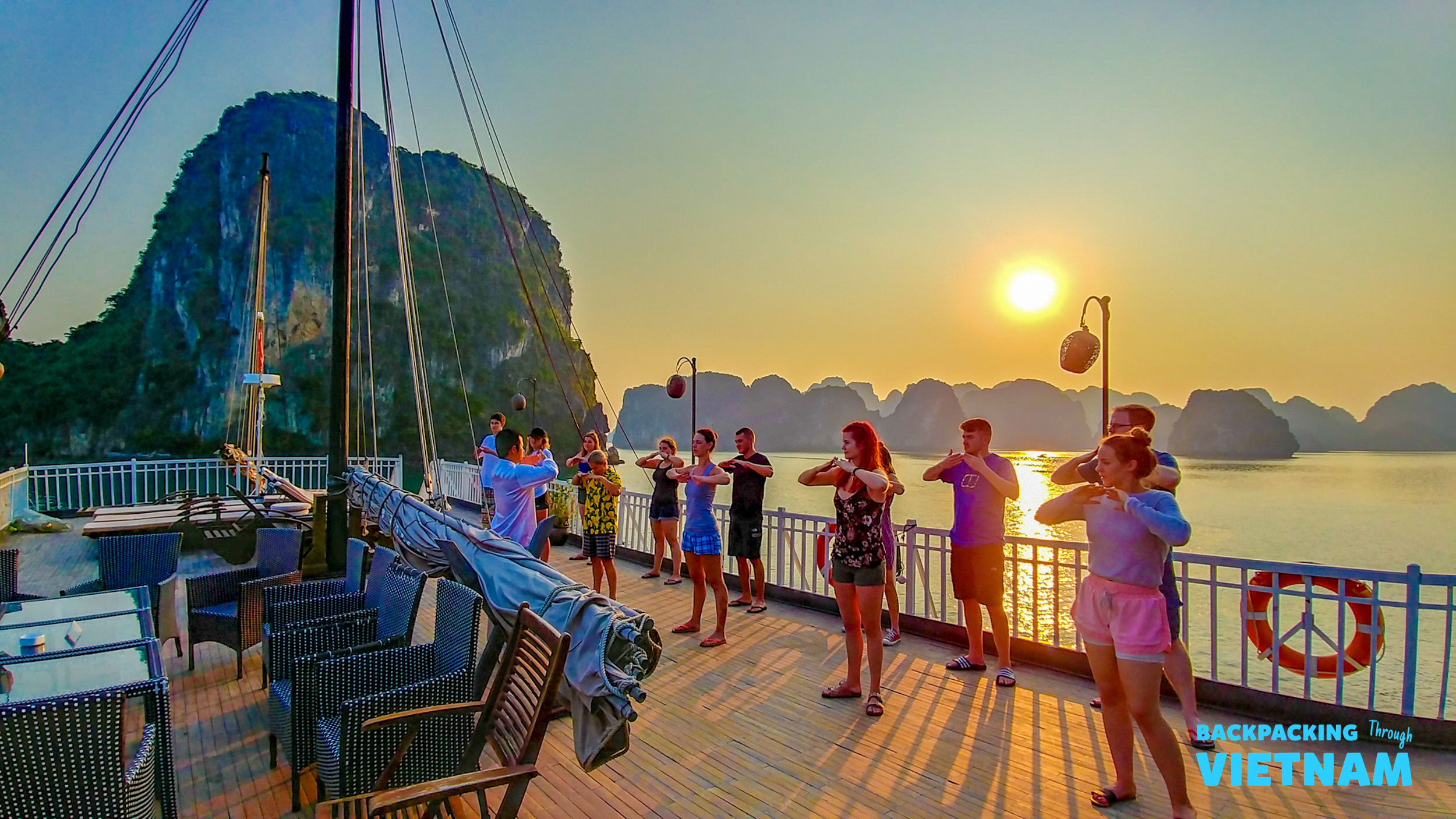Tourists practice tai chi on a boat deck at sunset, surrounded by limestone karsts in Halong Bay.
