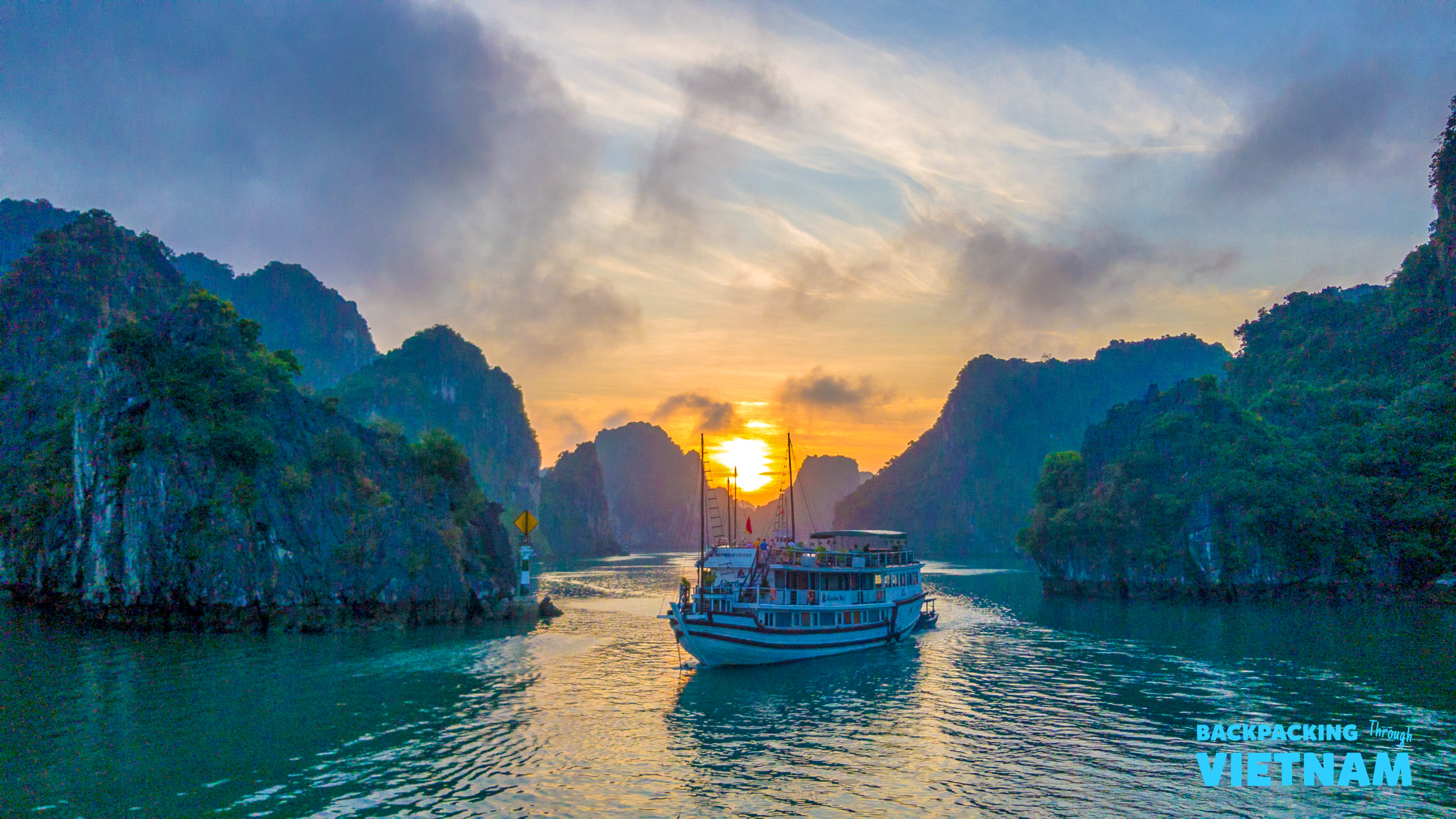 A tour boat cruises through the majestic limestone islands of Halong Bay at a beautiful sunset.