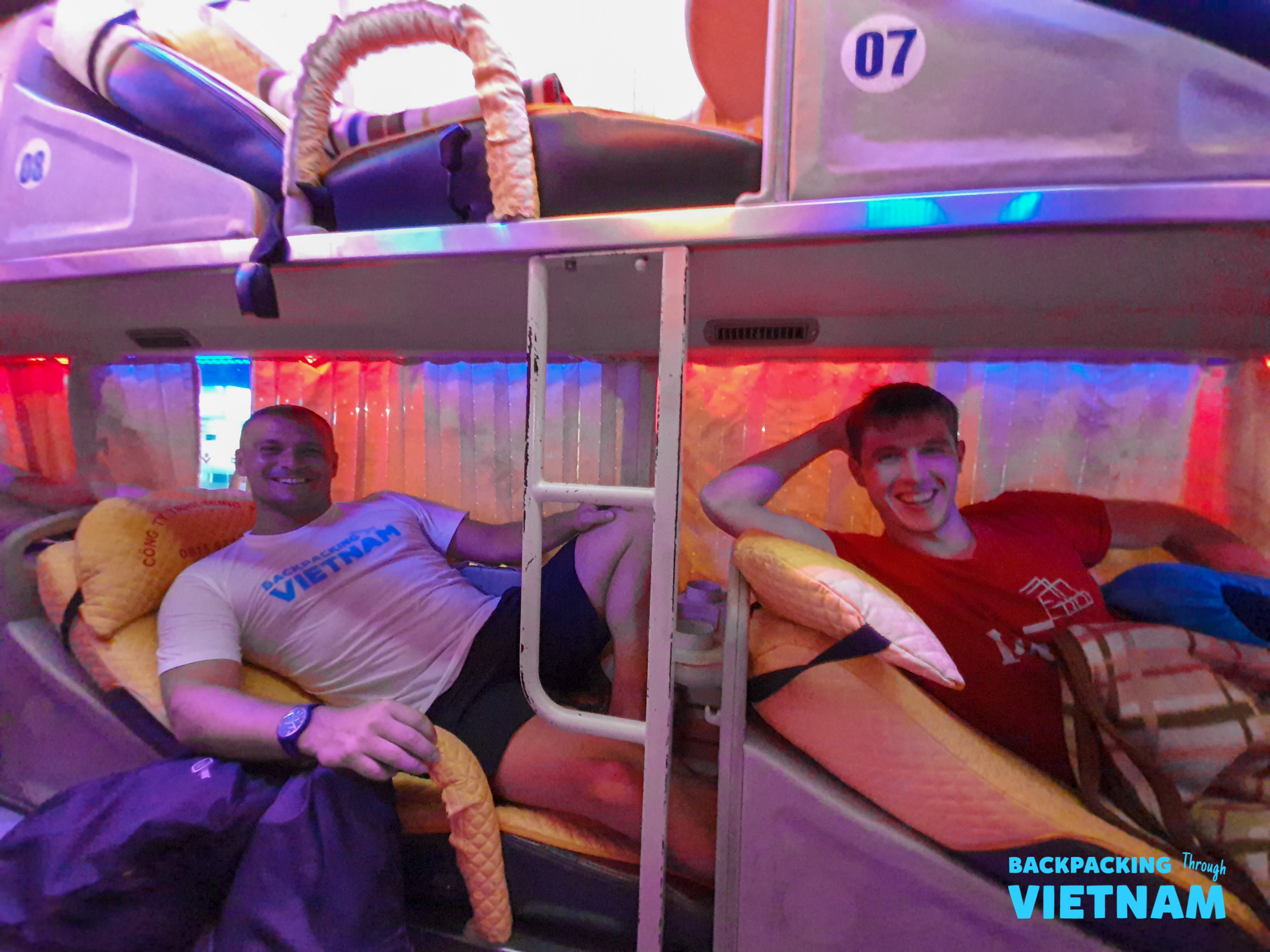 Two smiling men relax in their cozy, colorful bunks on a Vietnamese sleeper bus.