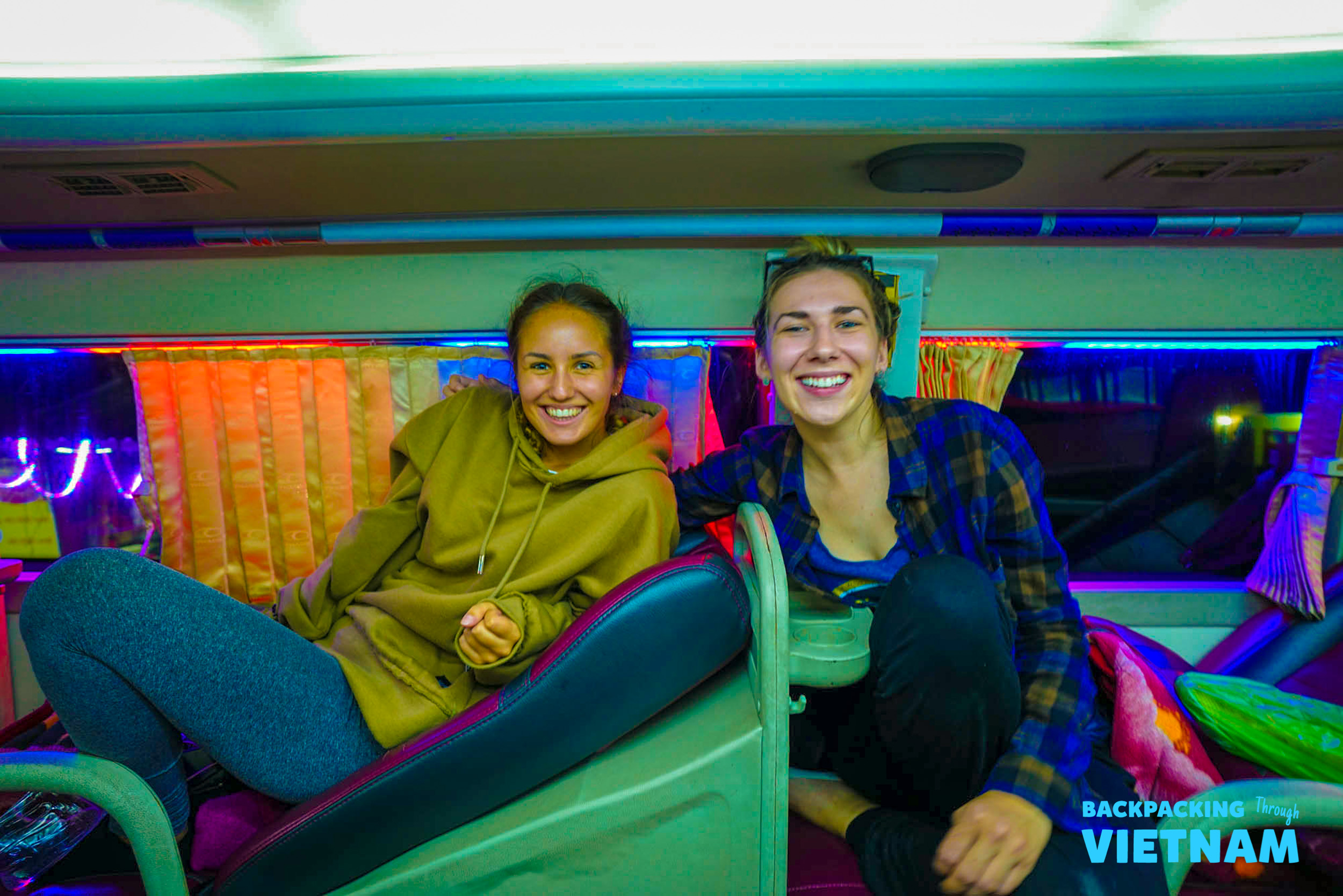 Two cheerful backpackers relaxing in a vibrant sleeper bus bunk on their Vietnam adventure.