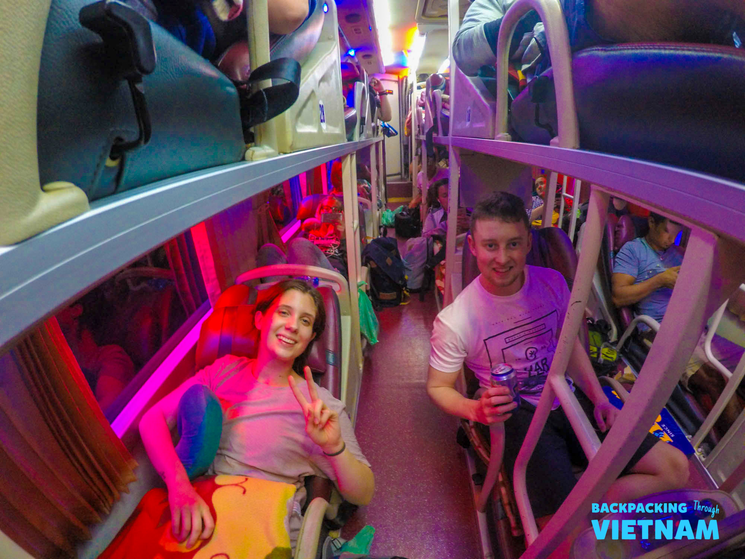 Two happy backpackers smiling from their bunks inside a colorful sleeper bus in Vietnam.