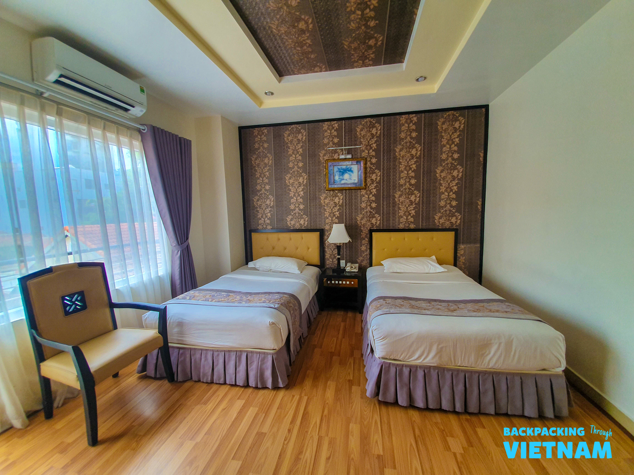A comfortable hotel room in Hue with two single beds, wooden floor, and patterned wallpaper.