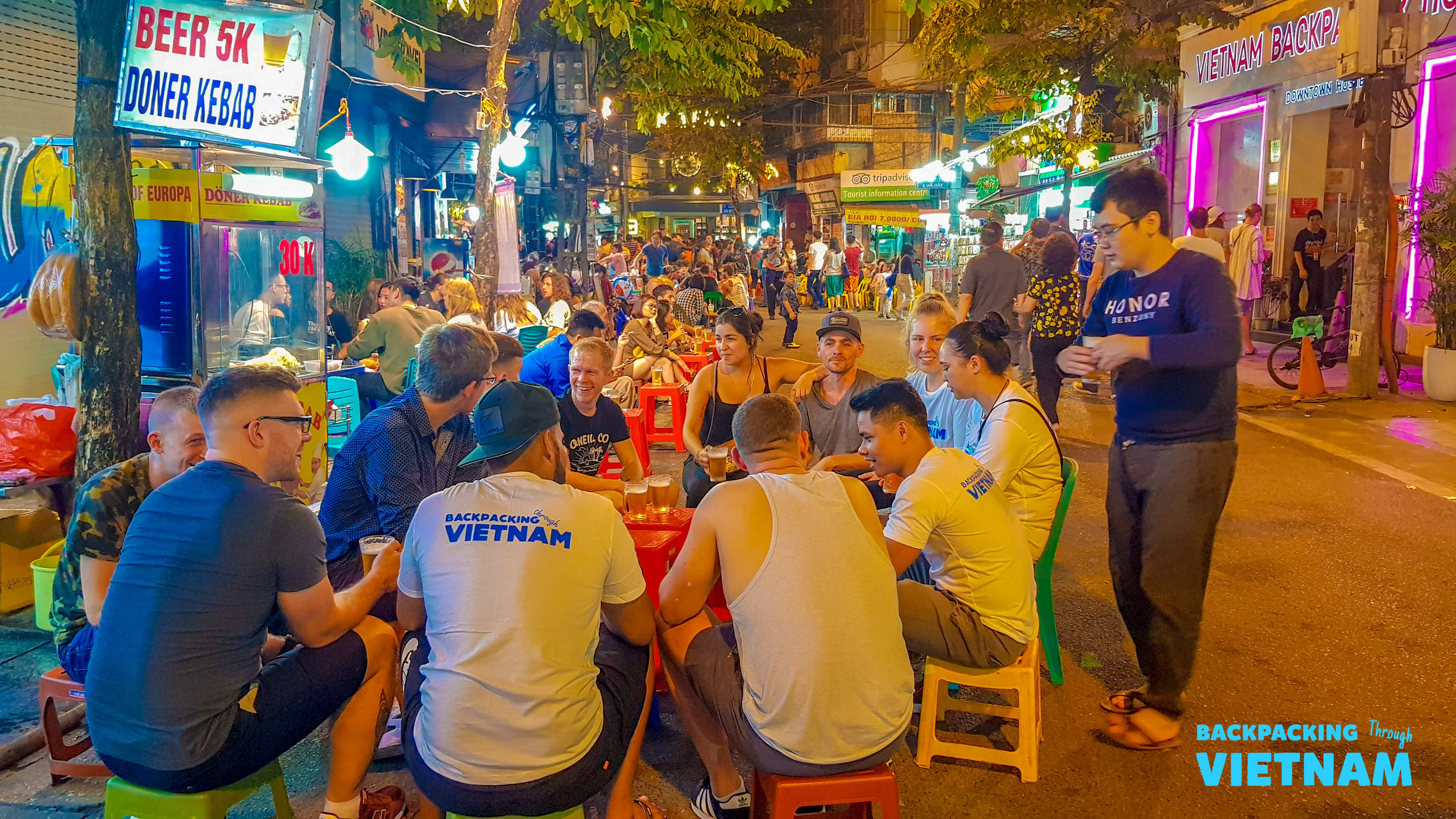 Backpackers enjoy a lively night out, drinking beer and socializing on a bustling street in Hanoi.