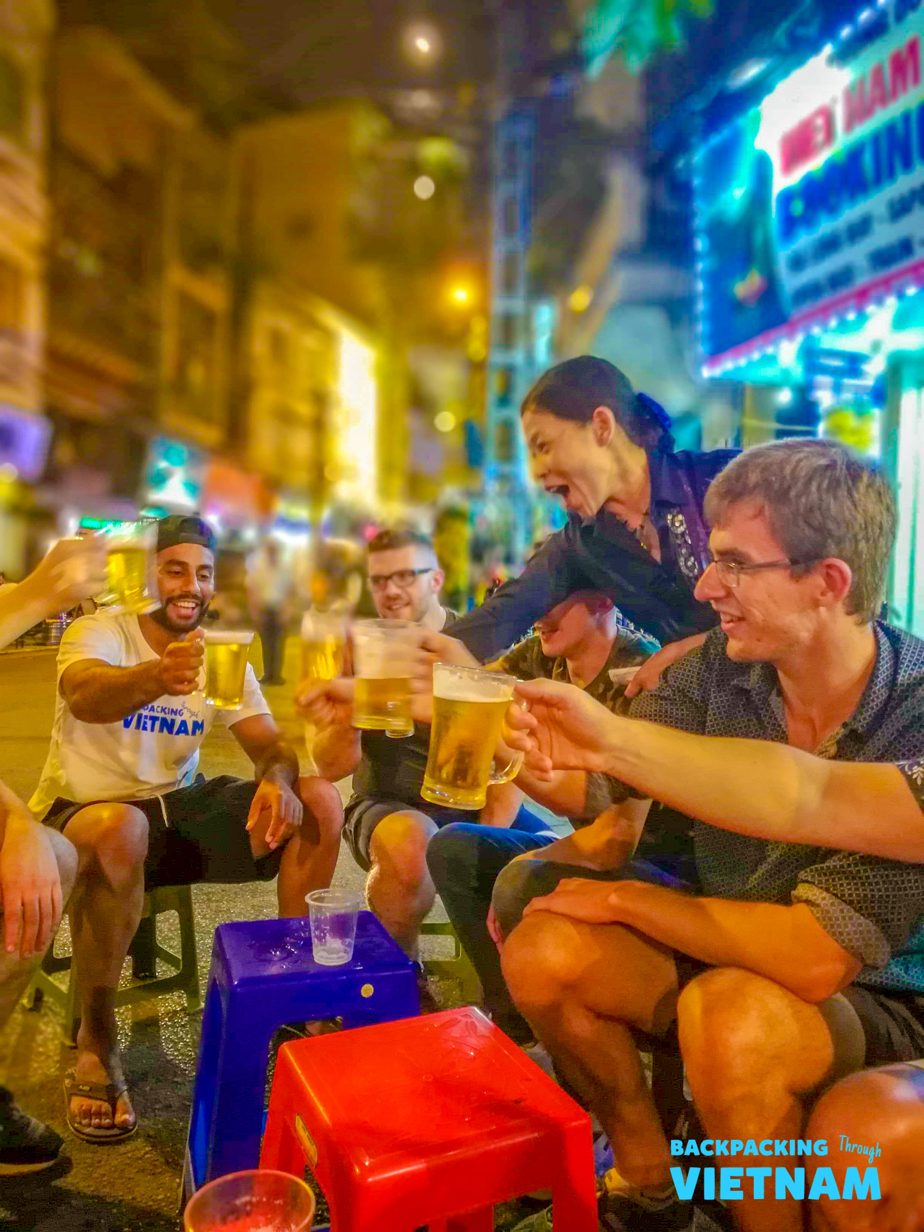 Smiling backpackers raise their beer glasses in a celebratory toast on a vibrant Hanoi street.