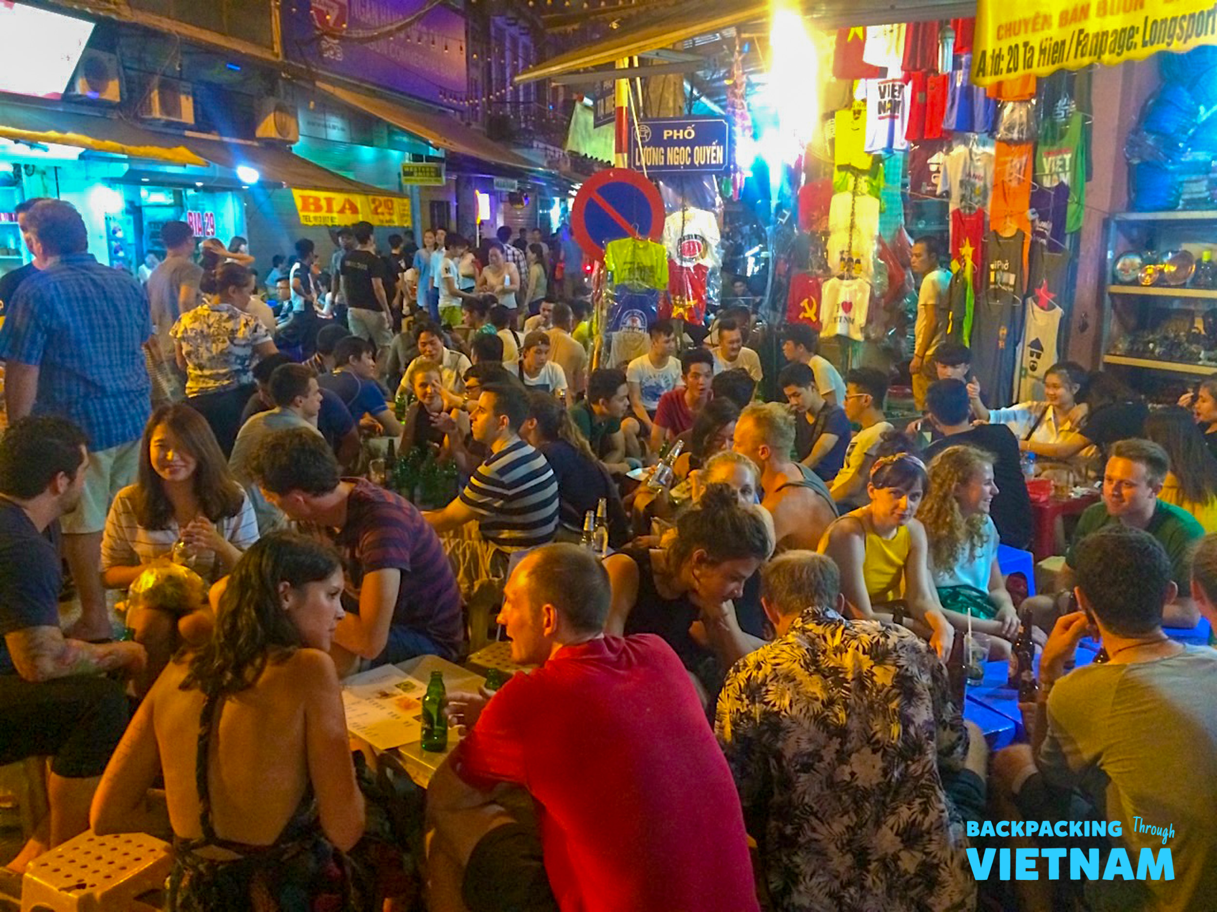 Backpackers gather on small stools, enjoying drinks on a vibrant, crowded street in Hanoi.