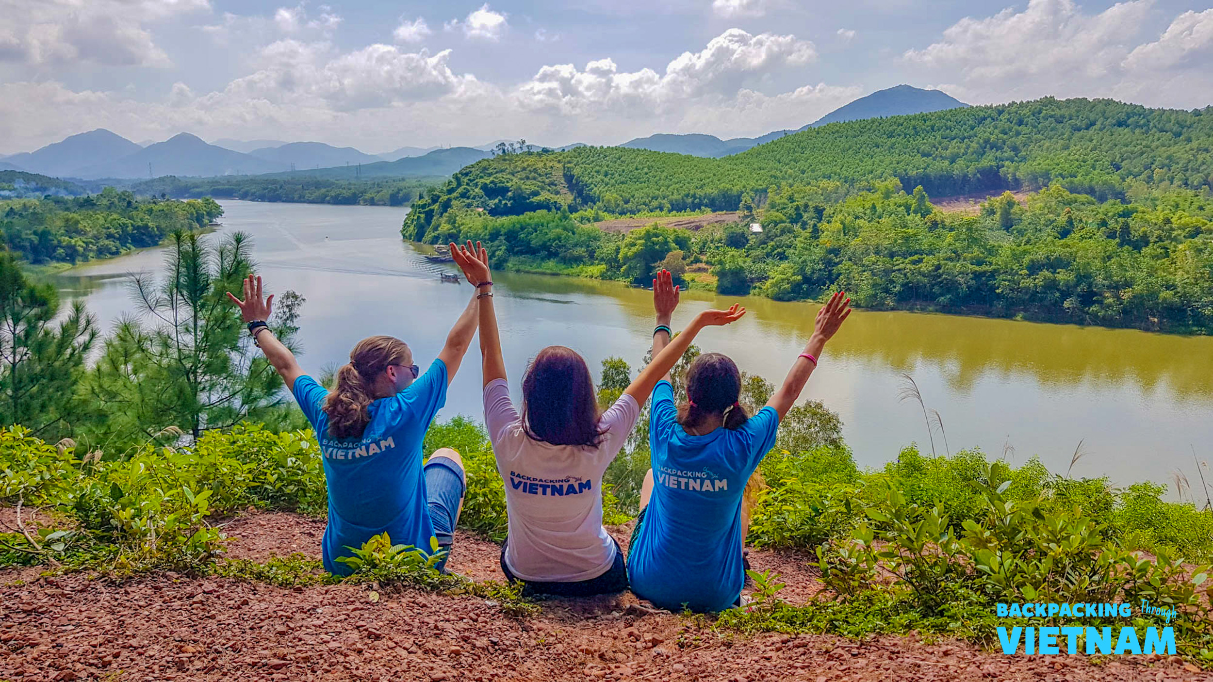Three friends with raised arms celebrating a beautiful river and mountain landscape in Vietnam.