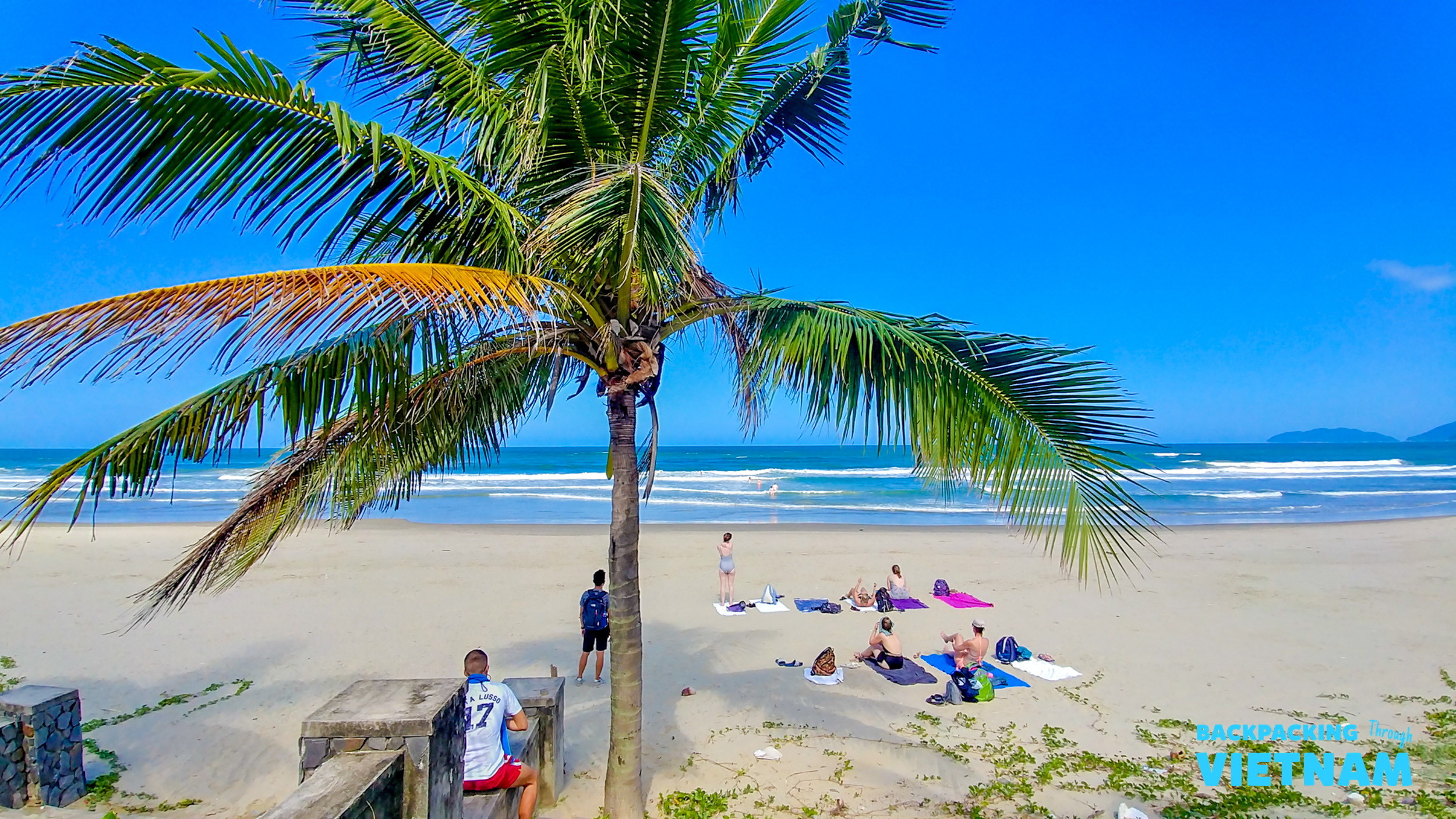 A tropical beach scene in Hoi An, Vietnam, with people relaxing under a palm tree.