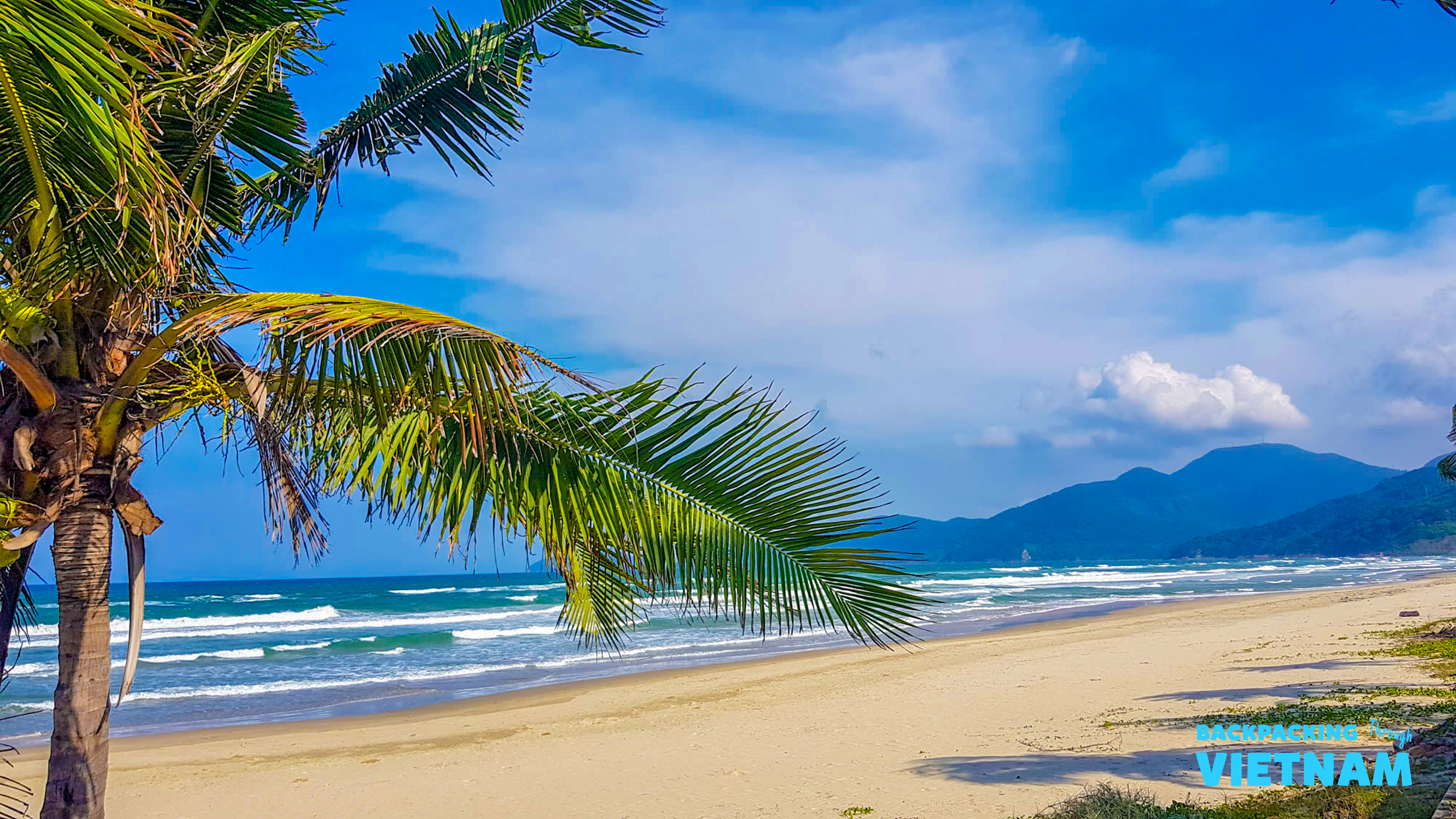 A sunny beach in Hoi An, Vietnam, with palm trees, ocean waves, and distant mountains.