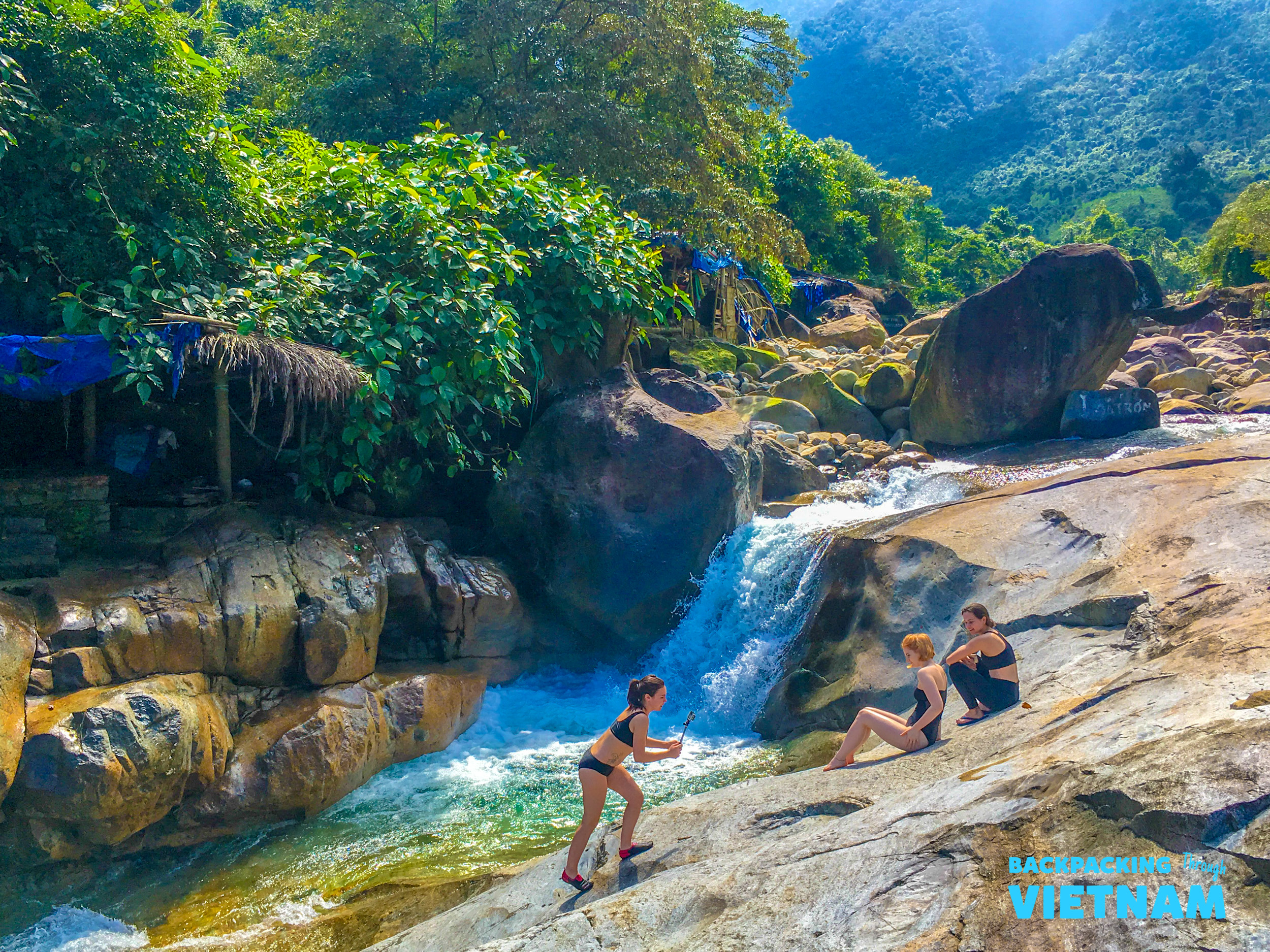 Three women relax and play in a beautiful natural spring with waterfalls and lush green mountains.