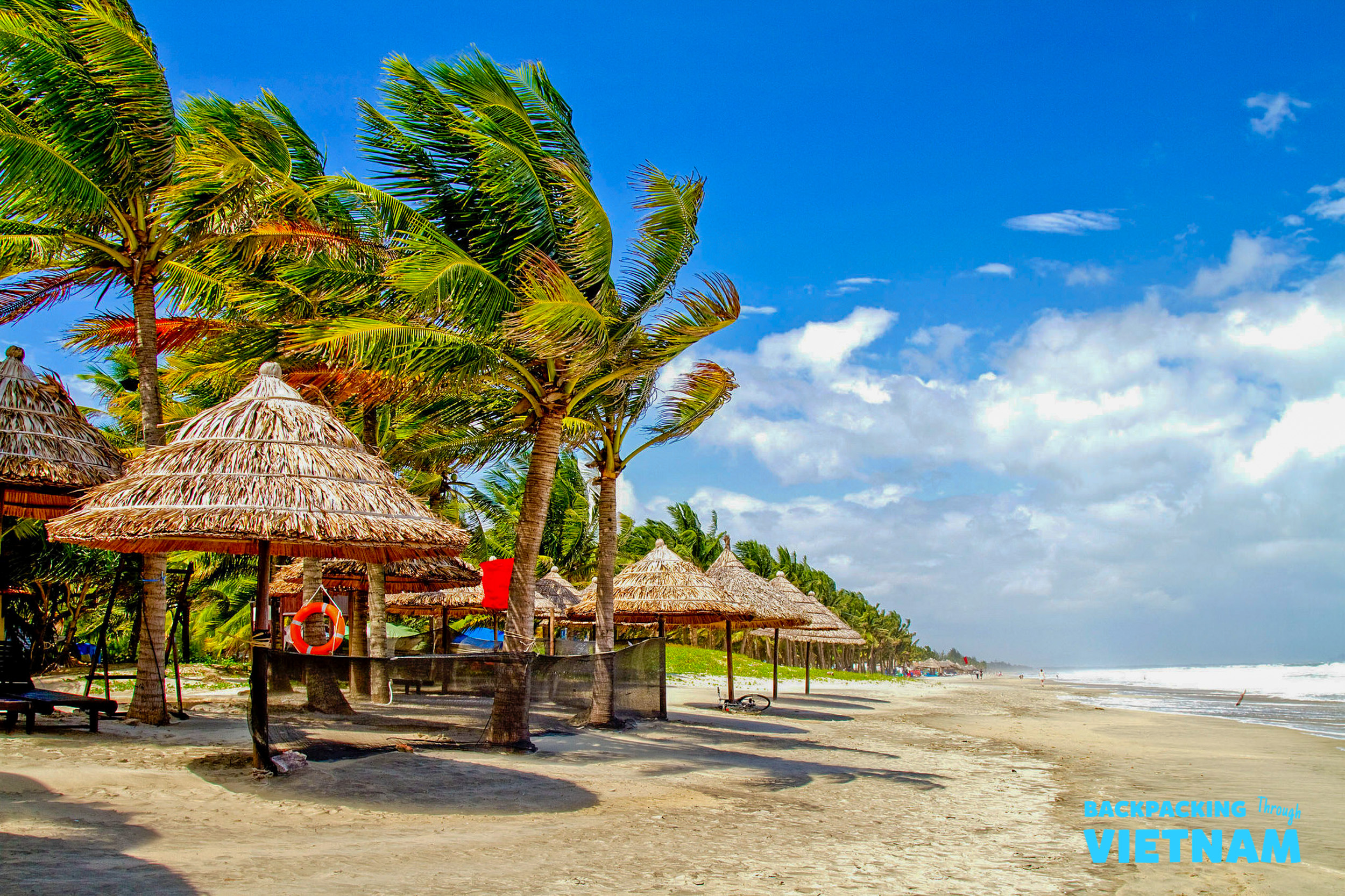 Sunny tropical beach with swaying palm trees and thatched umbrellas in Hoi An, Vietnam.