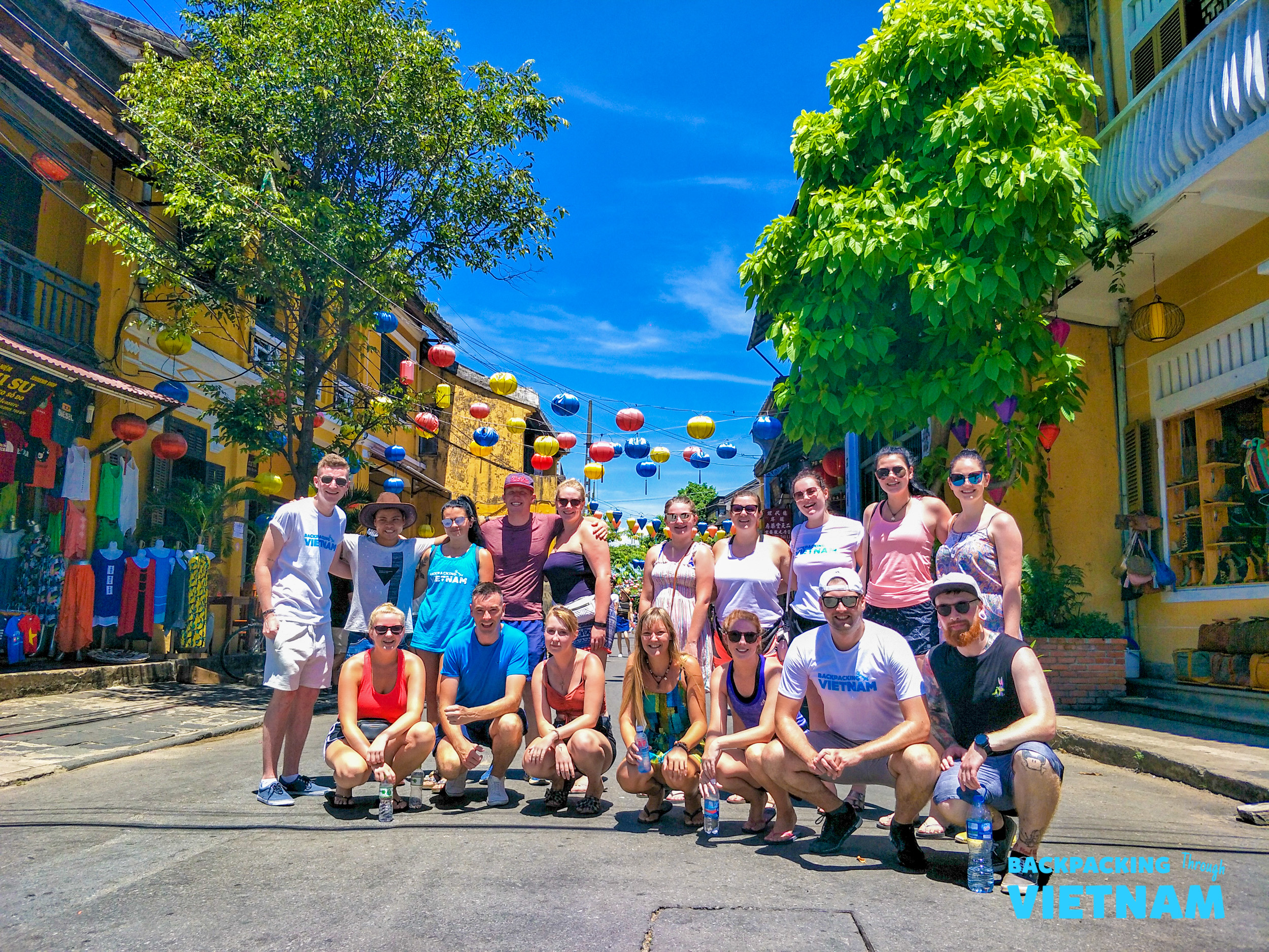 A group of happy backpackers posing on a sunny street in Hoi An, Vietnam, with colorful lanterns overhead.