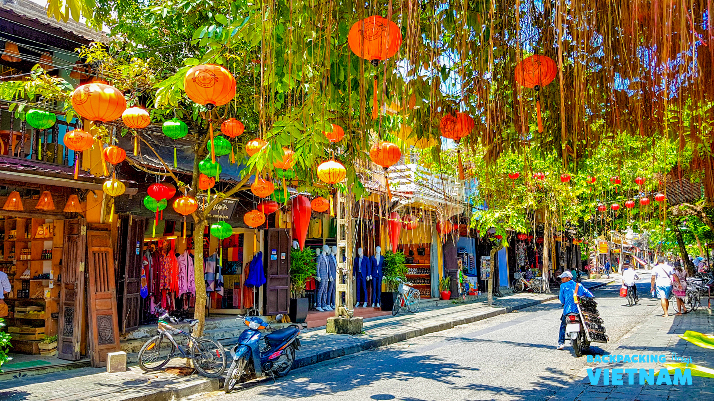 A vibrant street in Hoi An, Vietnam, bustling with people under colorful lanterns and lush trees.