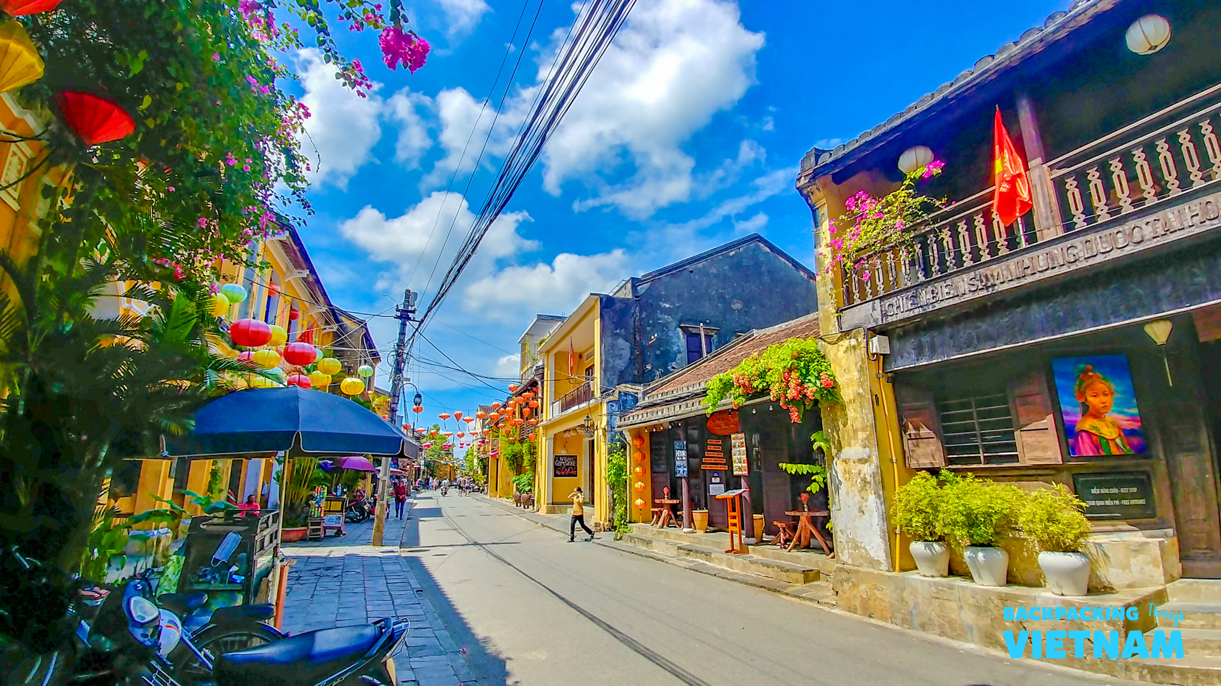 Colorful lanterns adorn historic buildings along a vibrant street in Hoi An, Vietnam, under a blue sky.