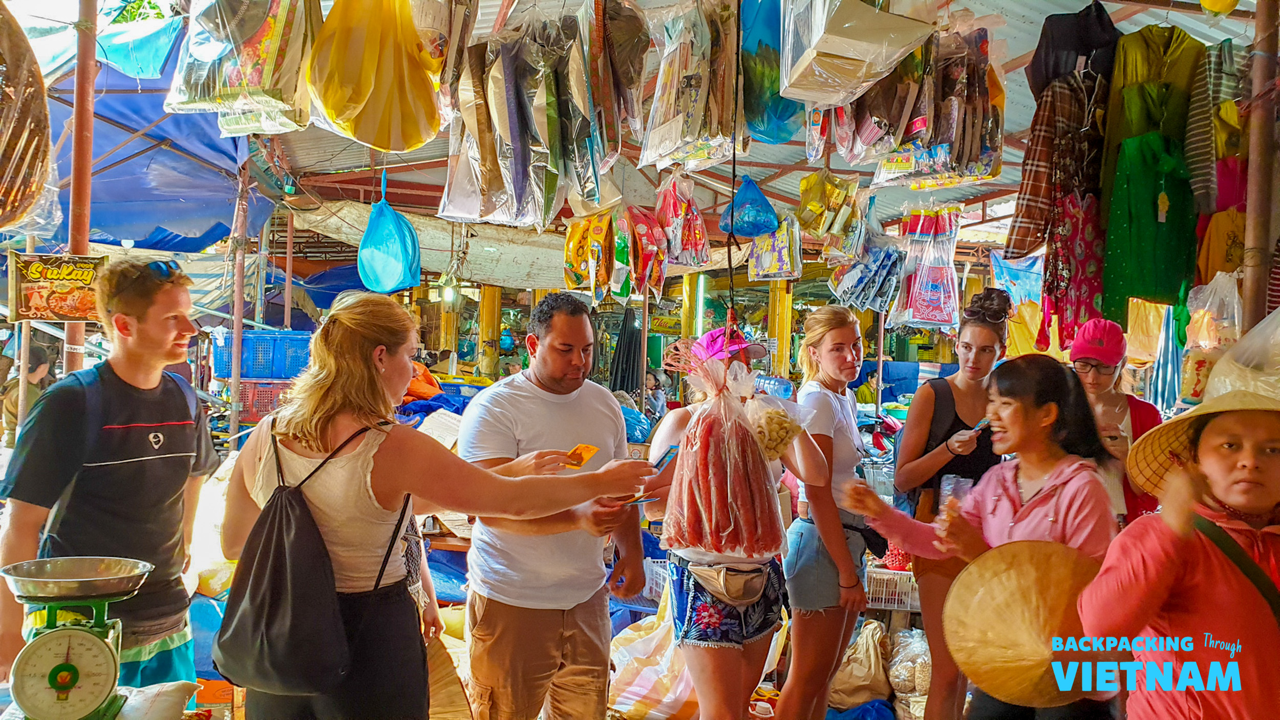 Backpackers explore a vibrant local market in Hoi An, interacting with vendors and browsing fresh produce.