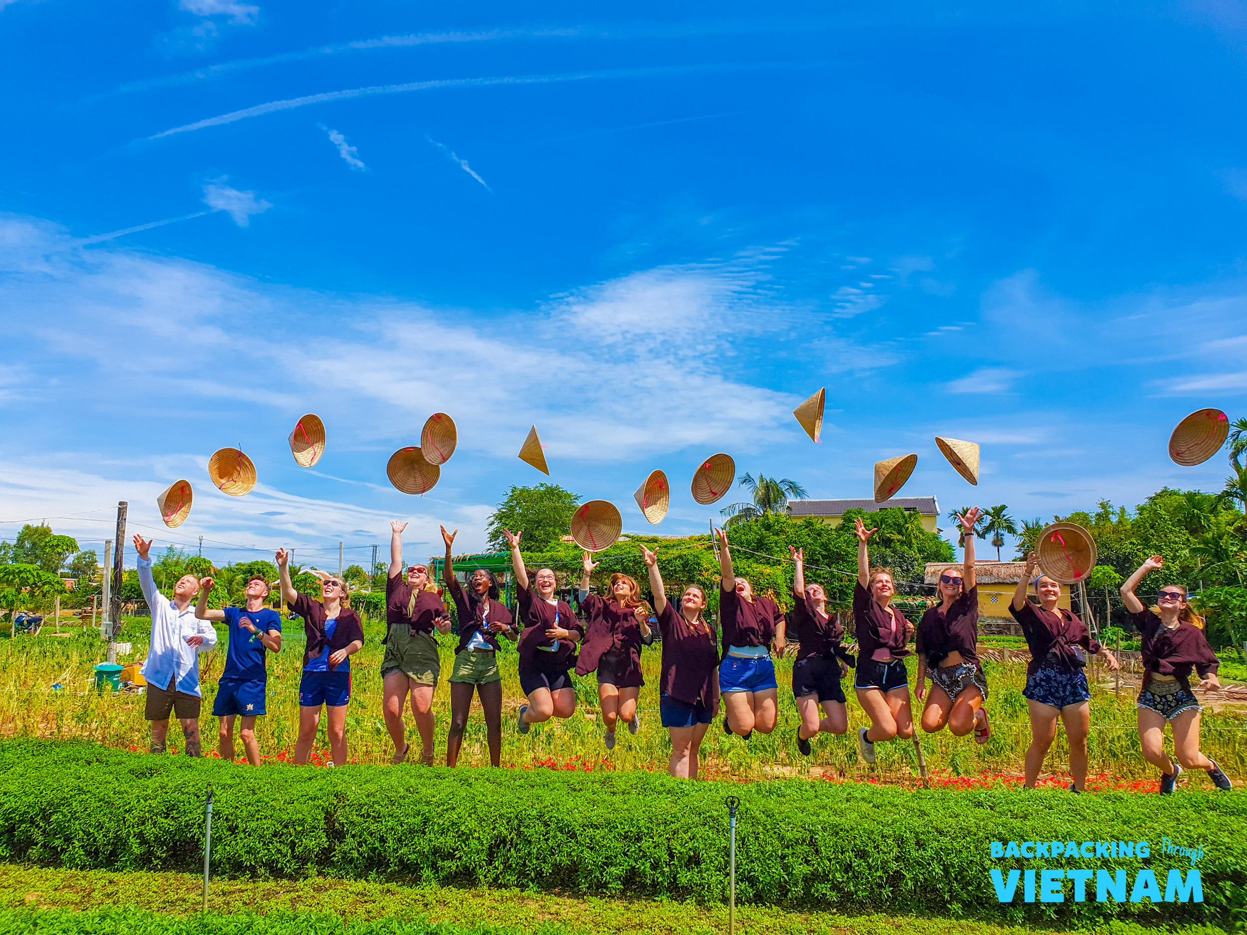 A joyful group of travelers jumping and throwing conical hats in a green field in Hoi An, Vietnam.