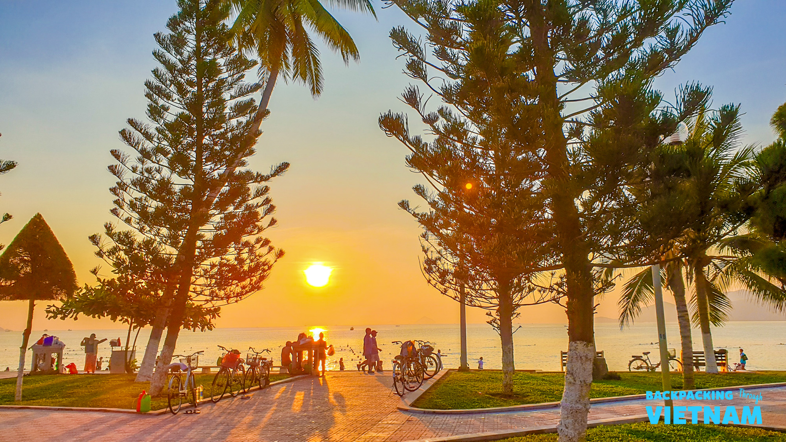 People gather along the Nha Trang coastline, enjoying a beautiful golden sunset over the ocean.