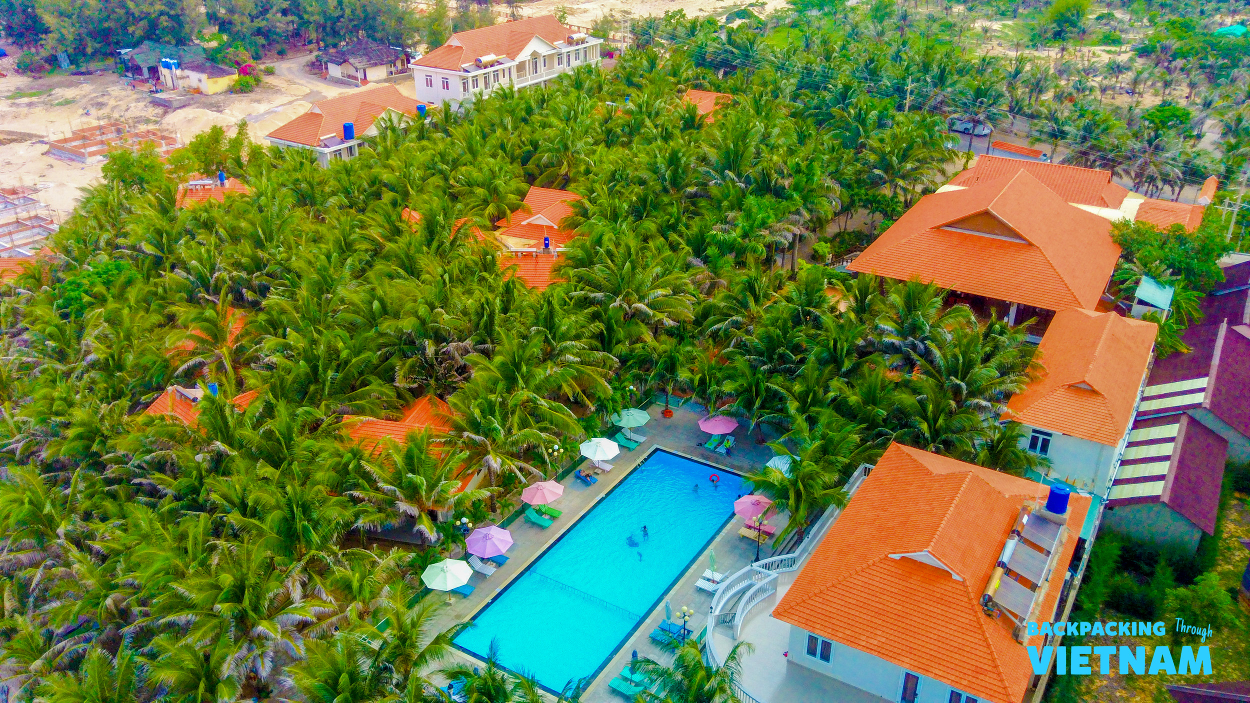 Vibrant Mui Ne resort with a long swimming pool, colorful umbrellas, and lush palm trees.
