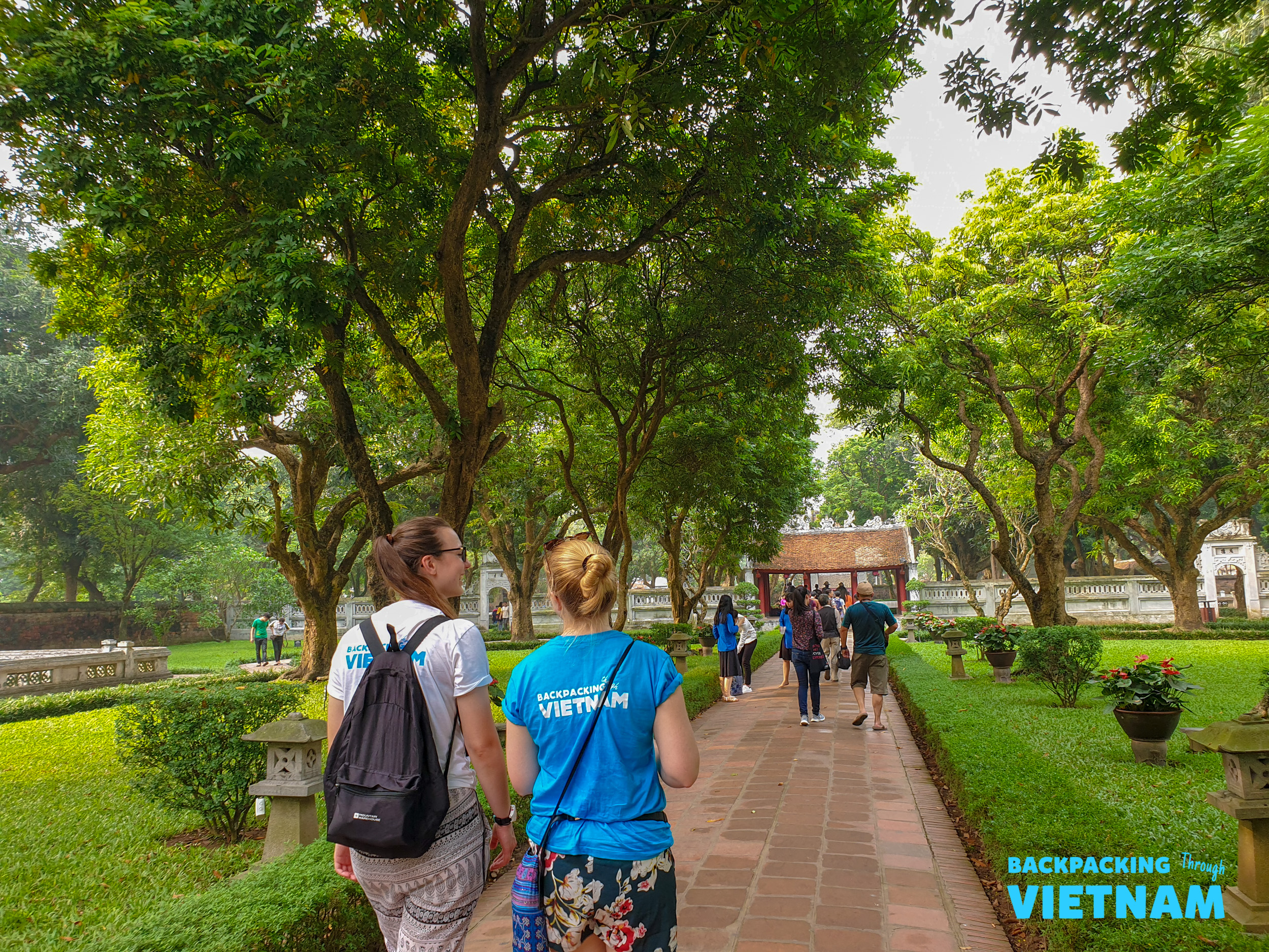 Two backpackers stroll through a beautiful, tree-lined garden with traditional architecture in Hanoi.