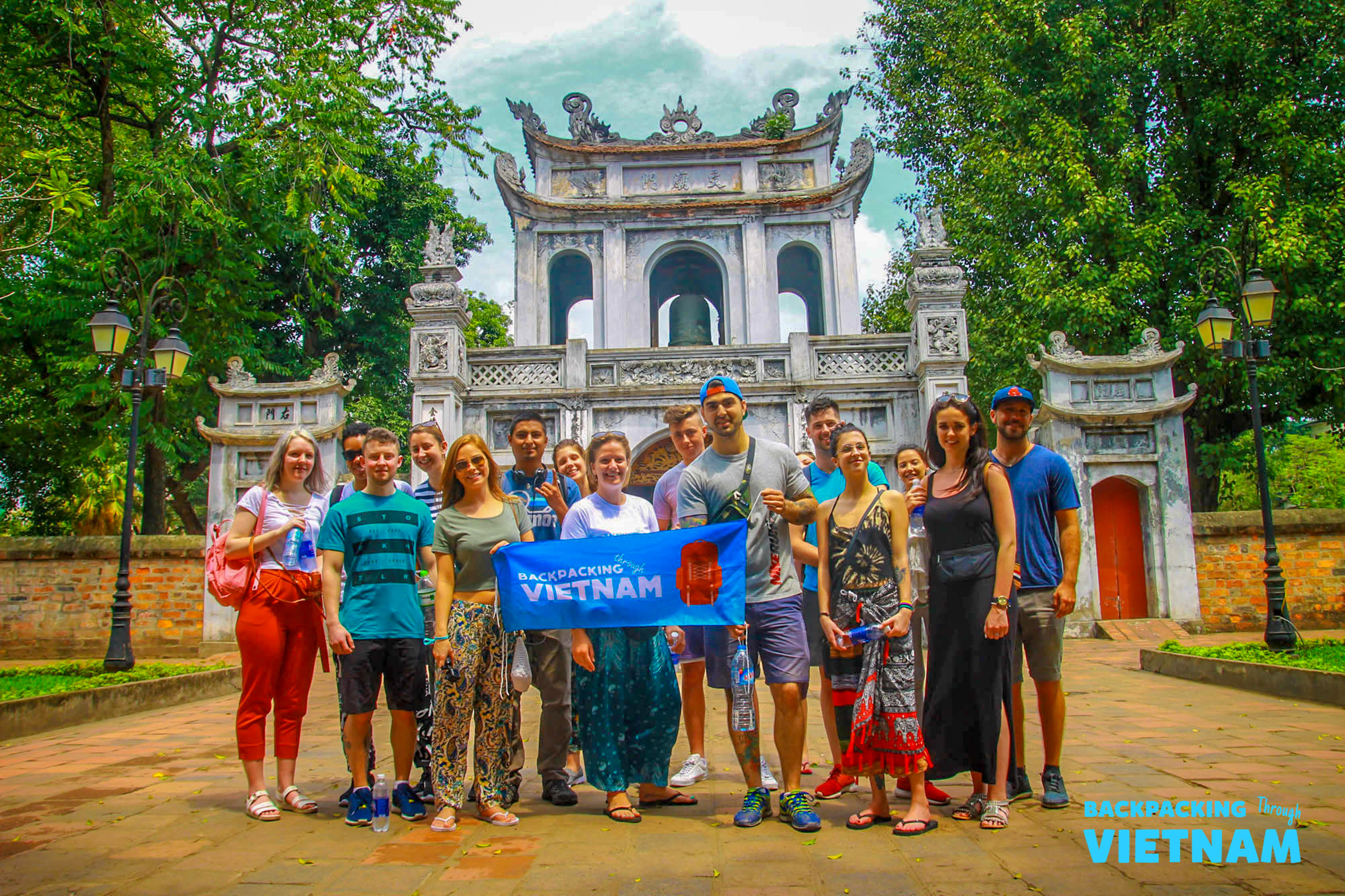 A group of happy backpackers posing with a 'Backpacking Vietnam' banner at a Hanoi temple.