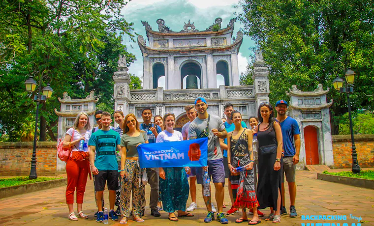 A group of happy backpackers posing with a 'Backpacking Vietnam' banner at a Hanoi temple.
