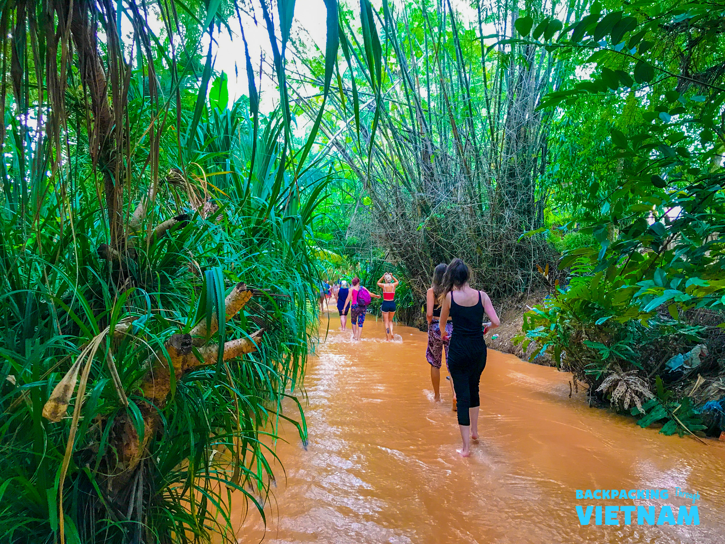 Backpackers walk barefoot through the shallow, reddish water of Vietnam's Fairy Stream, surrounded by dense jungle.