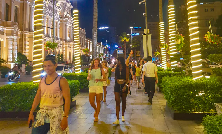 Friends enjoy a lively evening stroll past illuminated palm trees and historic buildings in Ho Chi Minh.