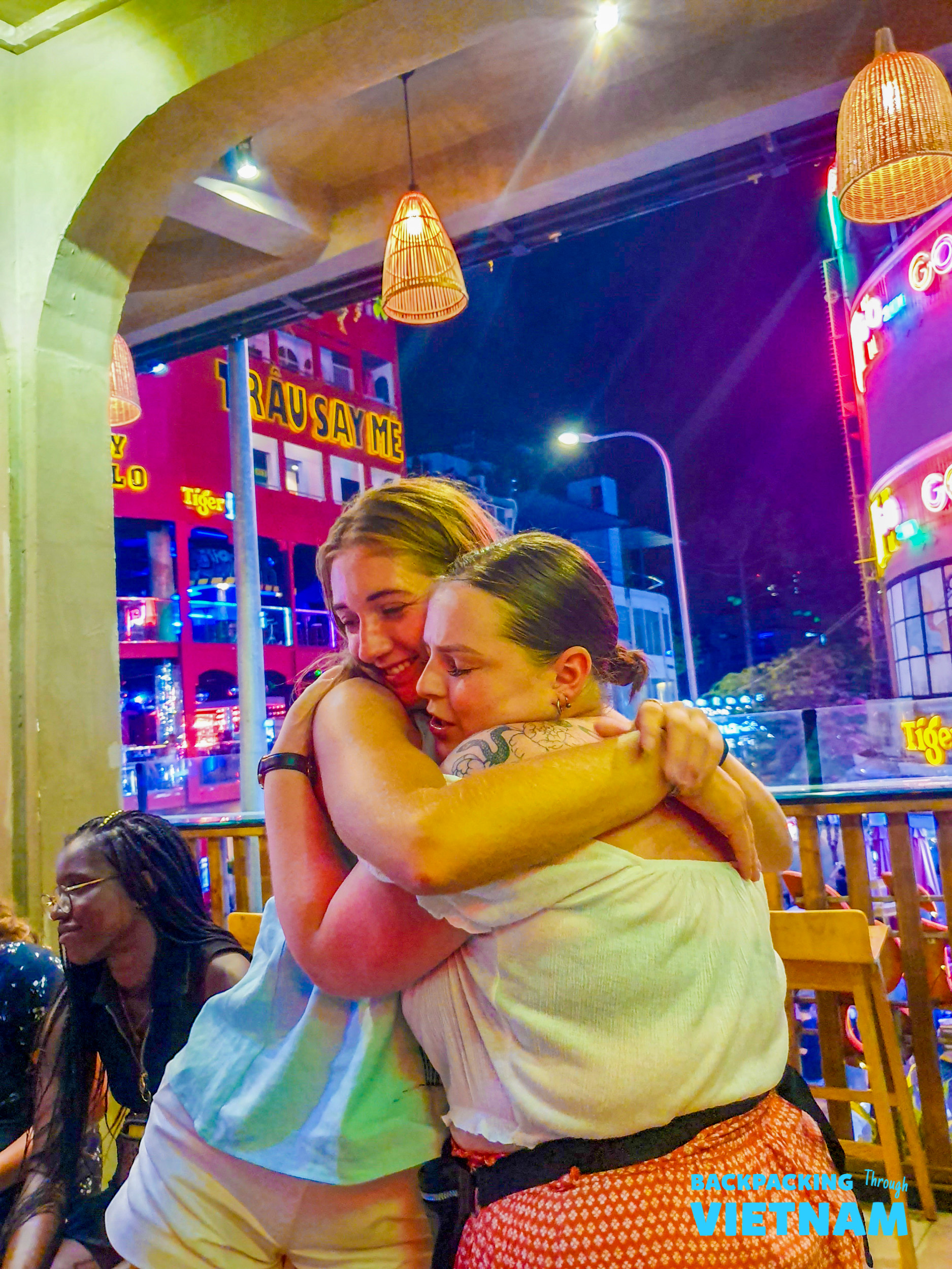 Two women share a heartfelt hug under neon lights during a lively night in Hoi An.