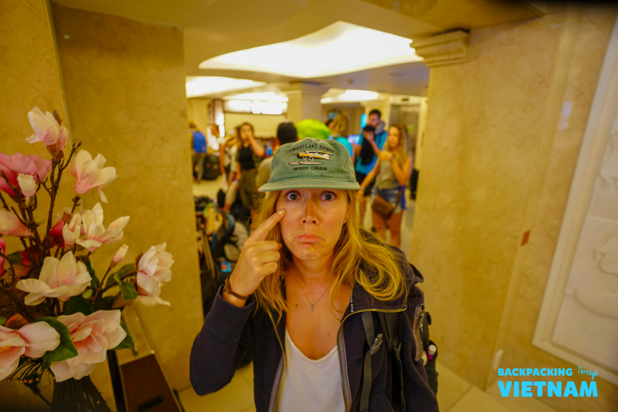 A backpacker makes a funny face, playfully reluctant to leave, surrounded by other travelers in Hoi An.