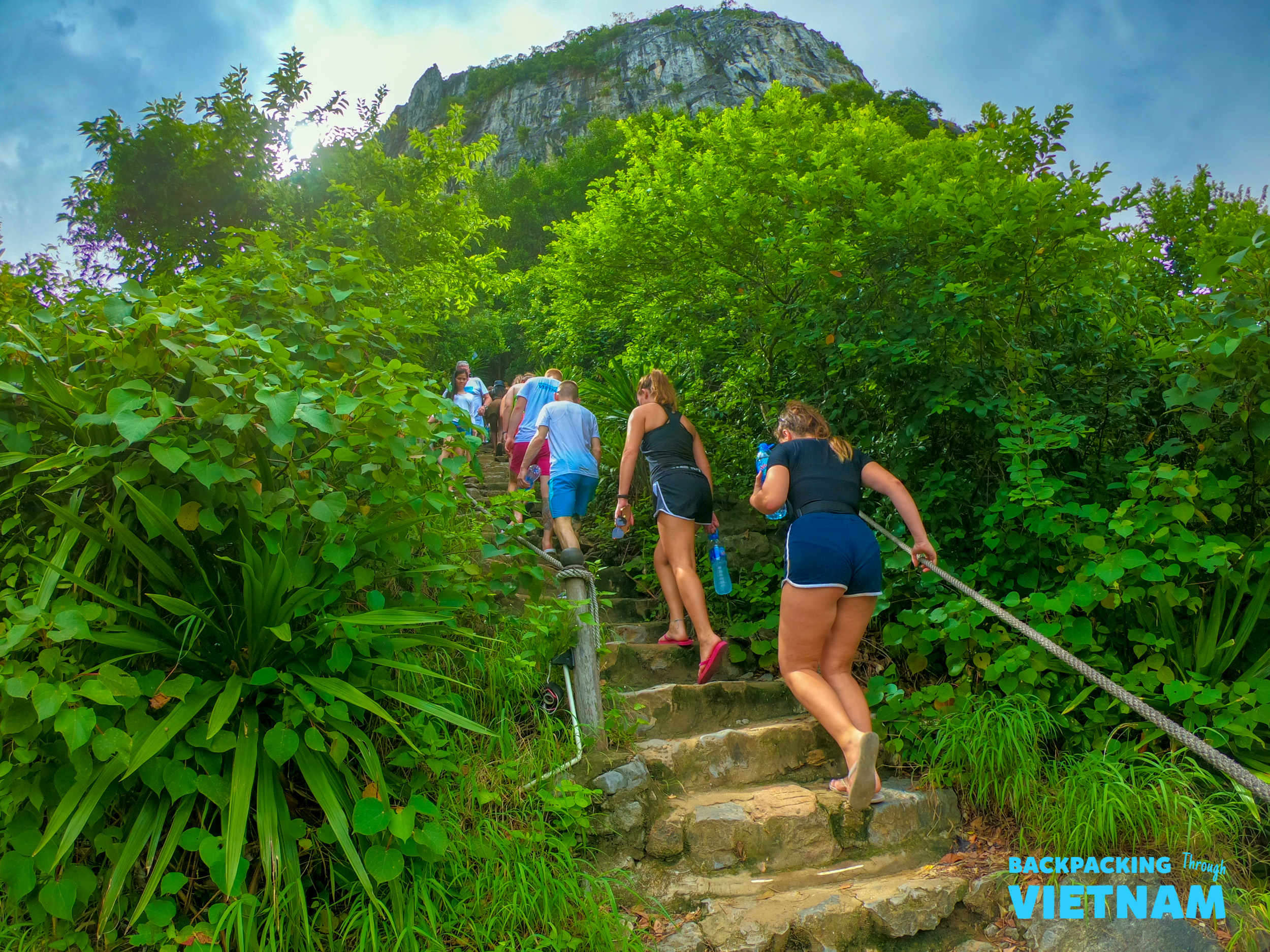 Backpackers climb steep stone steps surrounded by lush green foliage towards a rocky mountain in Tam Coc, Vietnam.