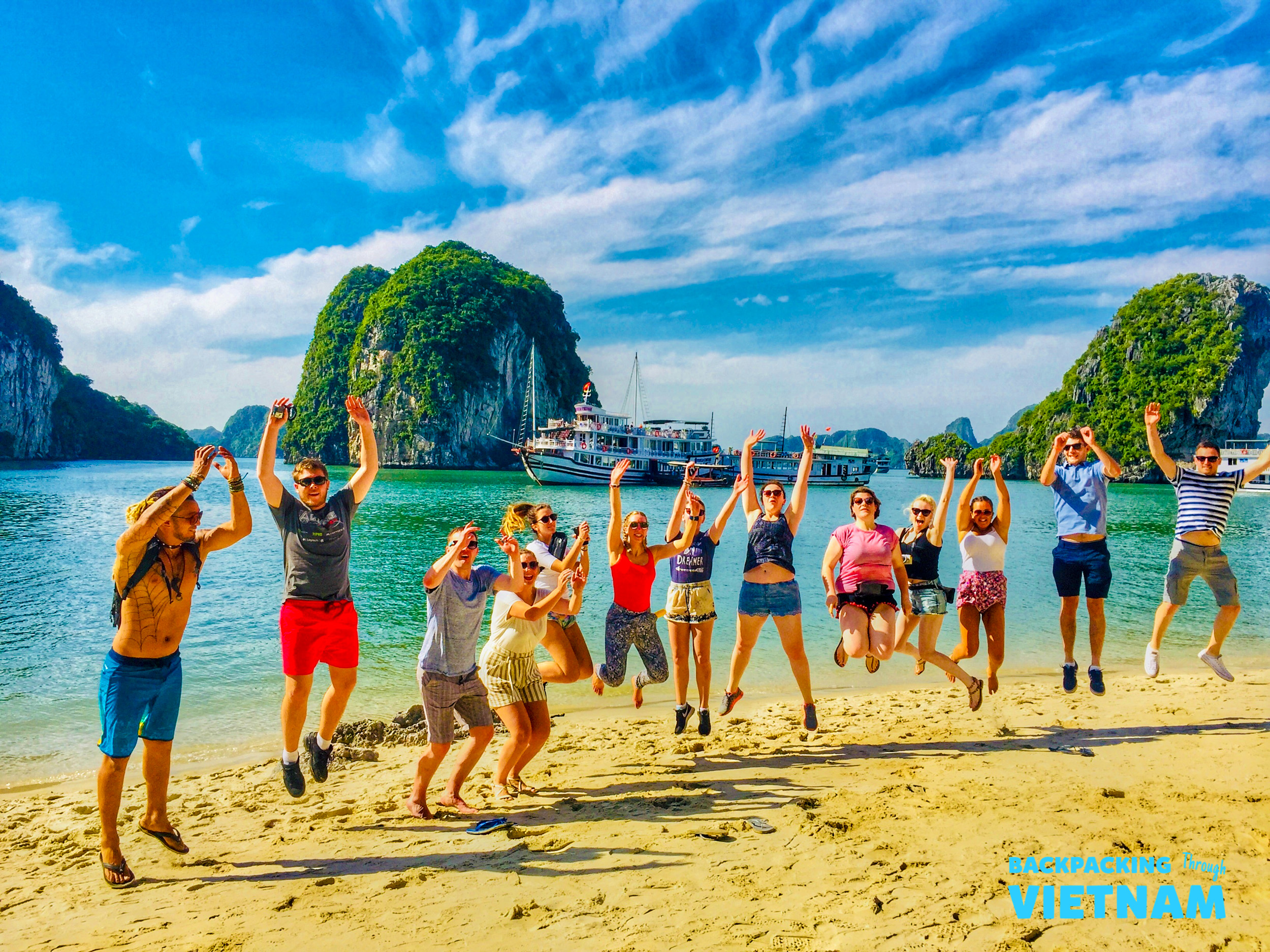 Happy backpackers jump on a sandy beach in Halong Bay, Vietnam, with stunning limestone karsts and a boat.