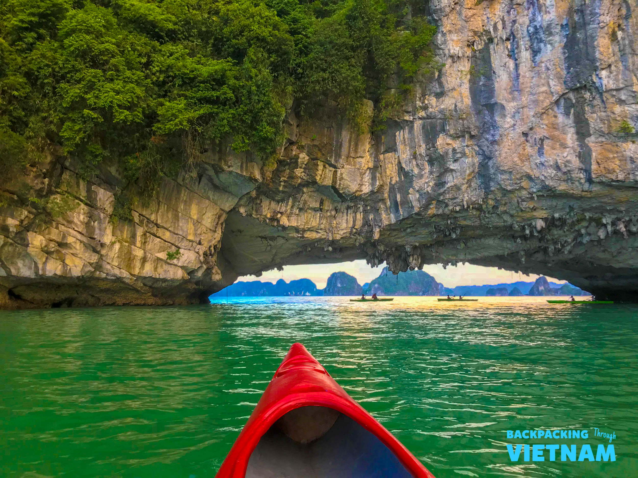 Kayaking through a stunning rock archway in Halong Bay, with other paddlers in the distance.